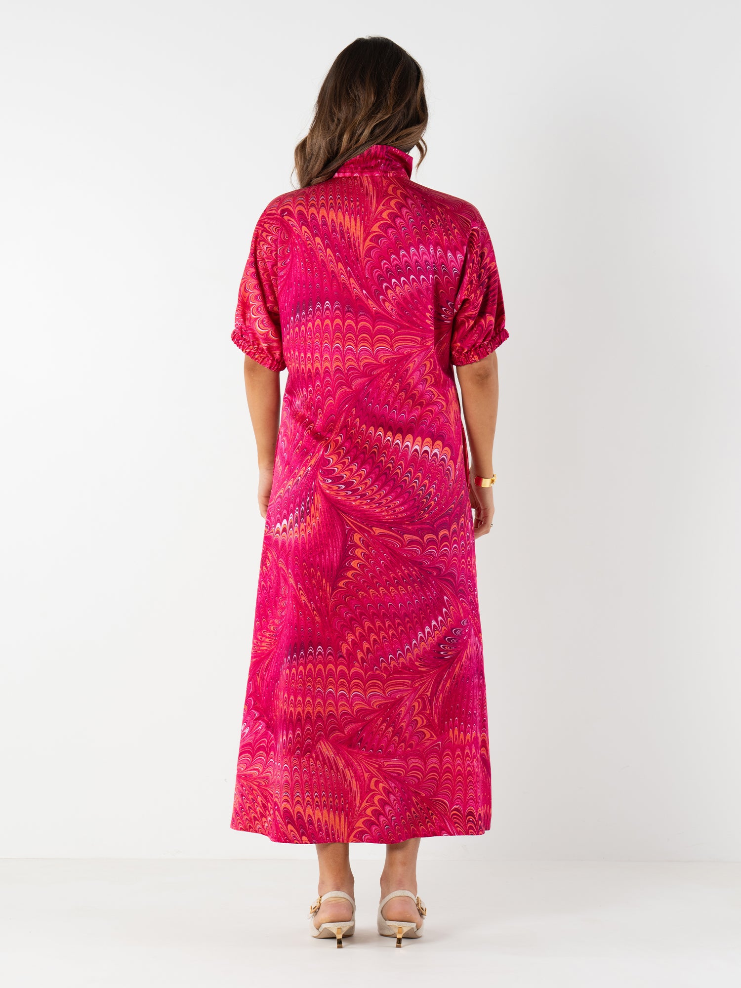 A woman with wavy brown hair and heeled sandals faces away, modeling the Poppy Maxi dress in a bright pink and red caftan silhouette against a plain white background.