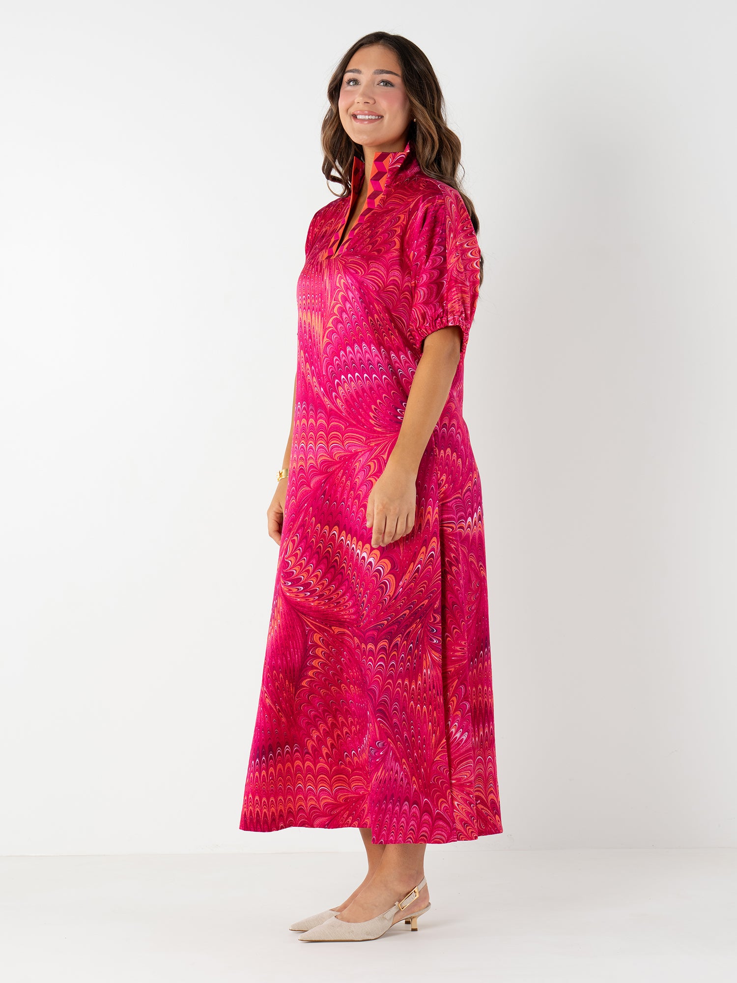 A woman with long brown hair smiles in the Poppy Maxi—an elegant bright pink Venetian print dress with short sleeves and a high collar. She pairs it with beige low-heeled shoes against a plain white background.