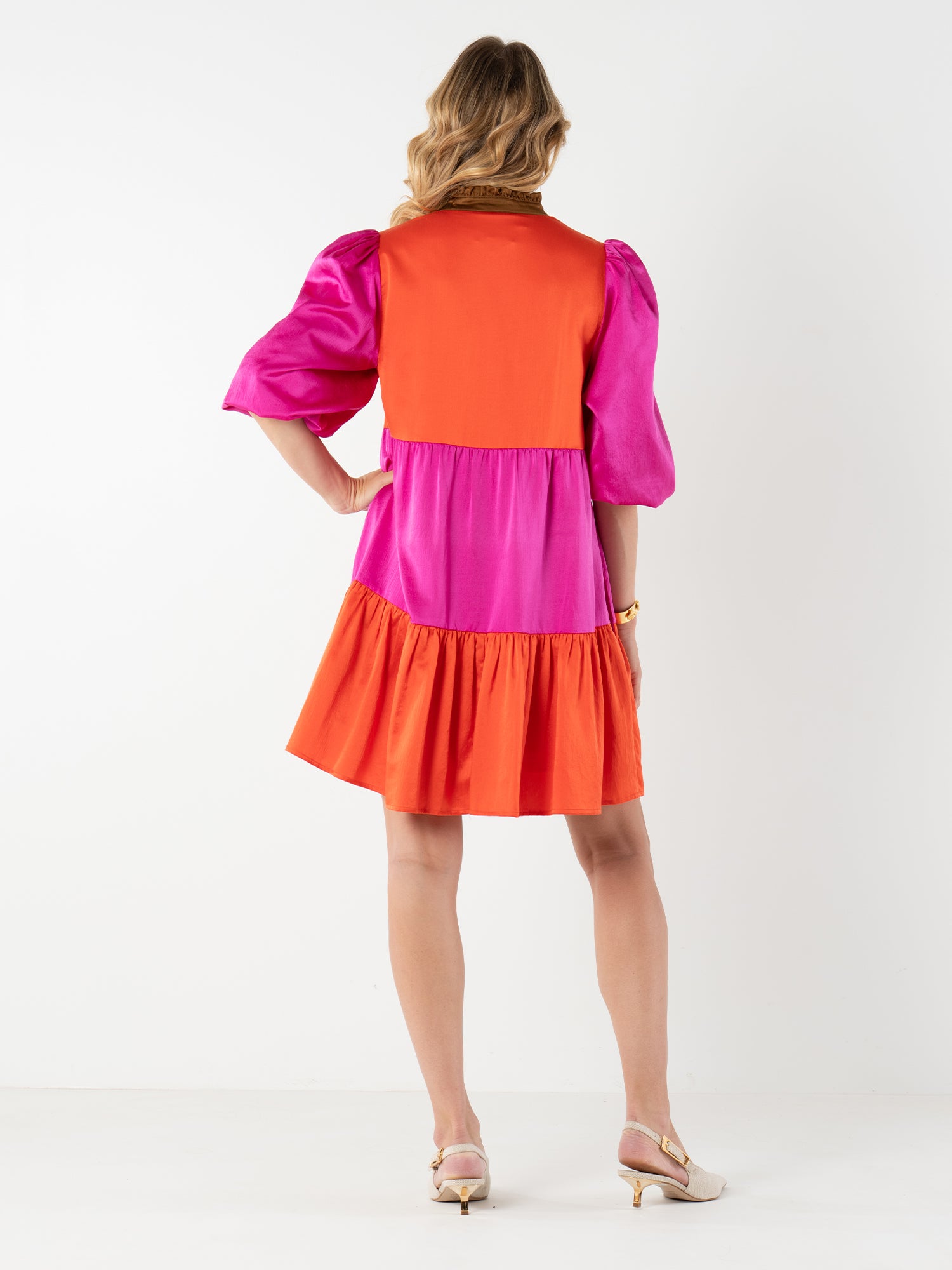 A woman with wavy blonde hair models the Sadie Dress—a color block dress with orange and magenta sections, puff sleeves, a tiered skirt, and mandarin collar—paired with cream heels against a white background.