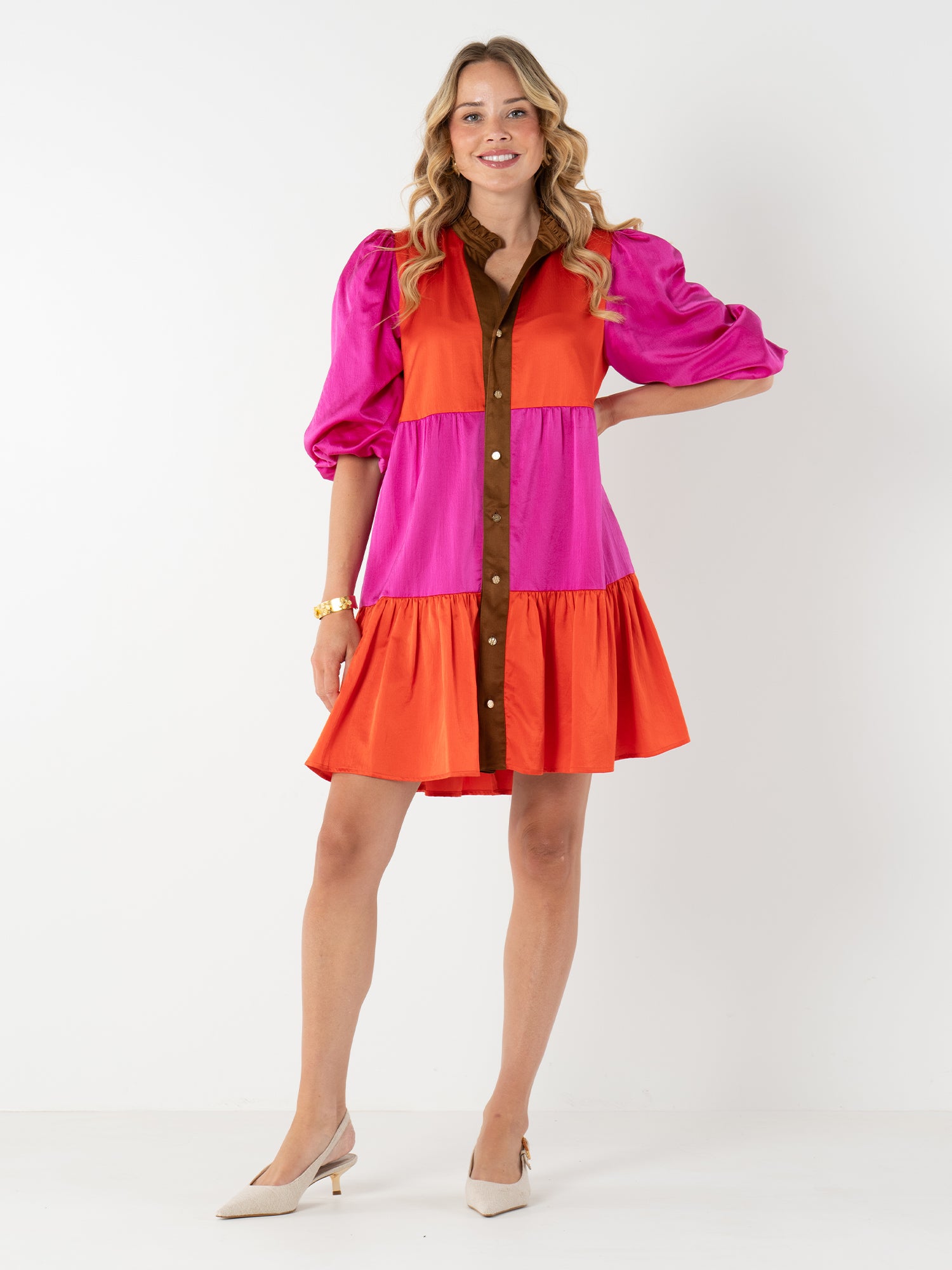A woman smiles in the Sadie Dress, featuring pink, orange, and brown color block panels, puffed sleeves, a mandarin collar, and front buttons. She pairs it with beige heels and a gold bracelet against a white background.