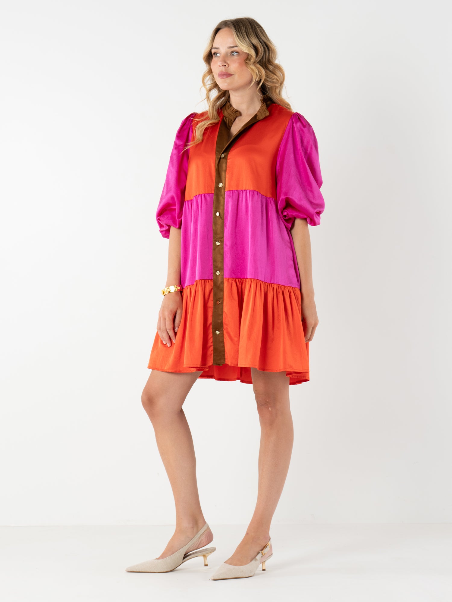A woman models the Sadie Dress, a loose color block dress with pink, orange, and red panels, brown button front, mandarin collar, and puff sleeves. She pairs it with light heels and a bracelet against a white background.