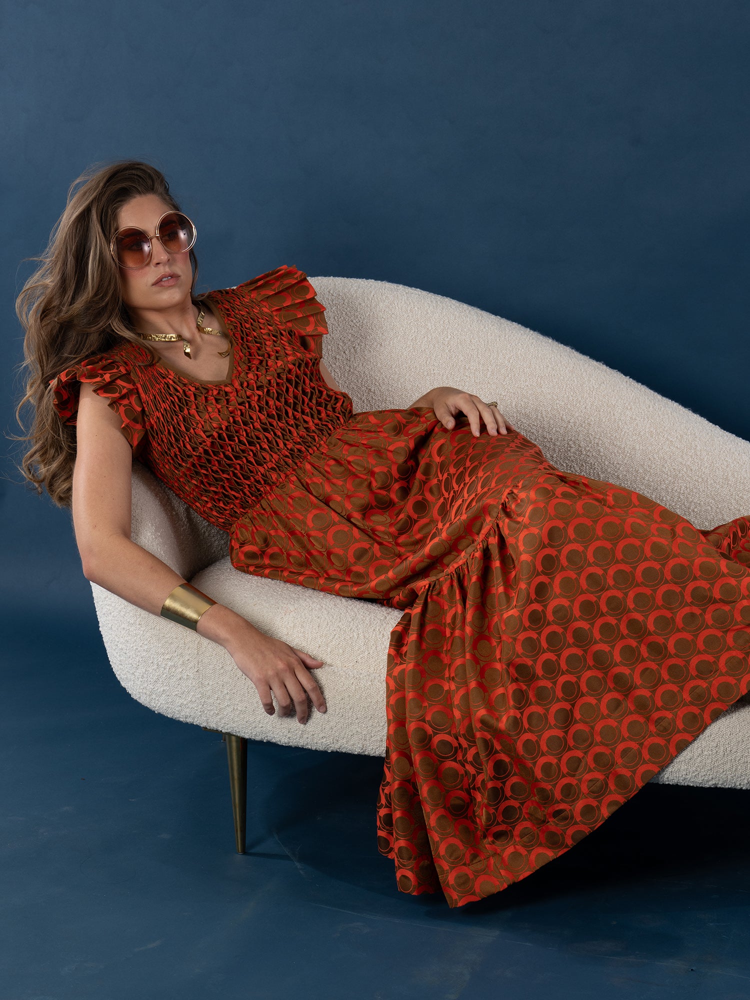 A woman in the Grace Maxi dress lounges on a white curved sofa, wearing round sunglasses, wavy hair, and gold jewelry, set against a dark blue background.