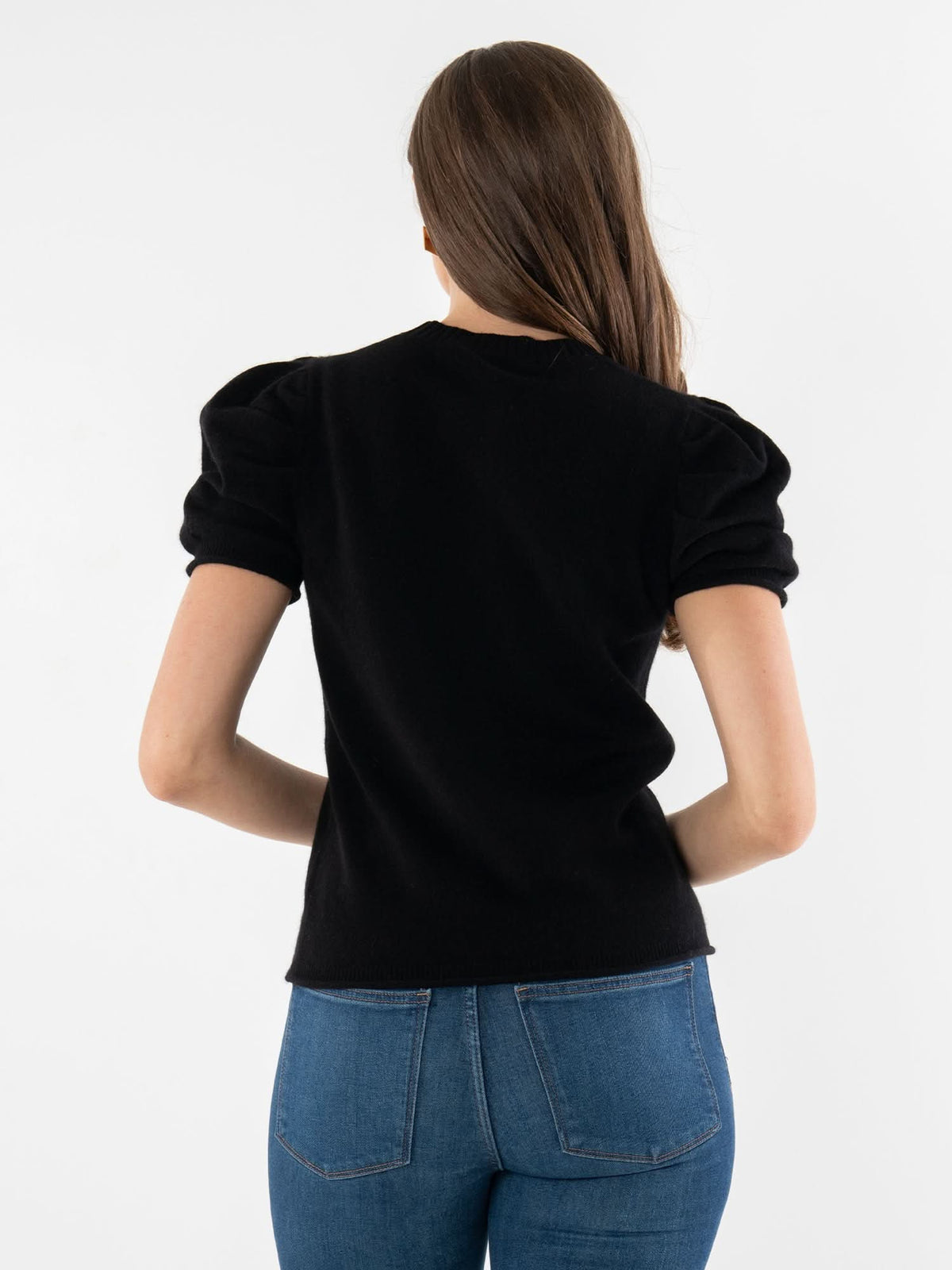 A woman with long brown hair stands with her back to the camera, wearing a luxury Crewneck Tee with puff sleeves and blue jeans, set against a plain white background.