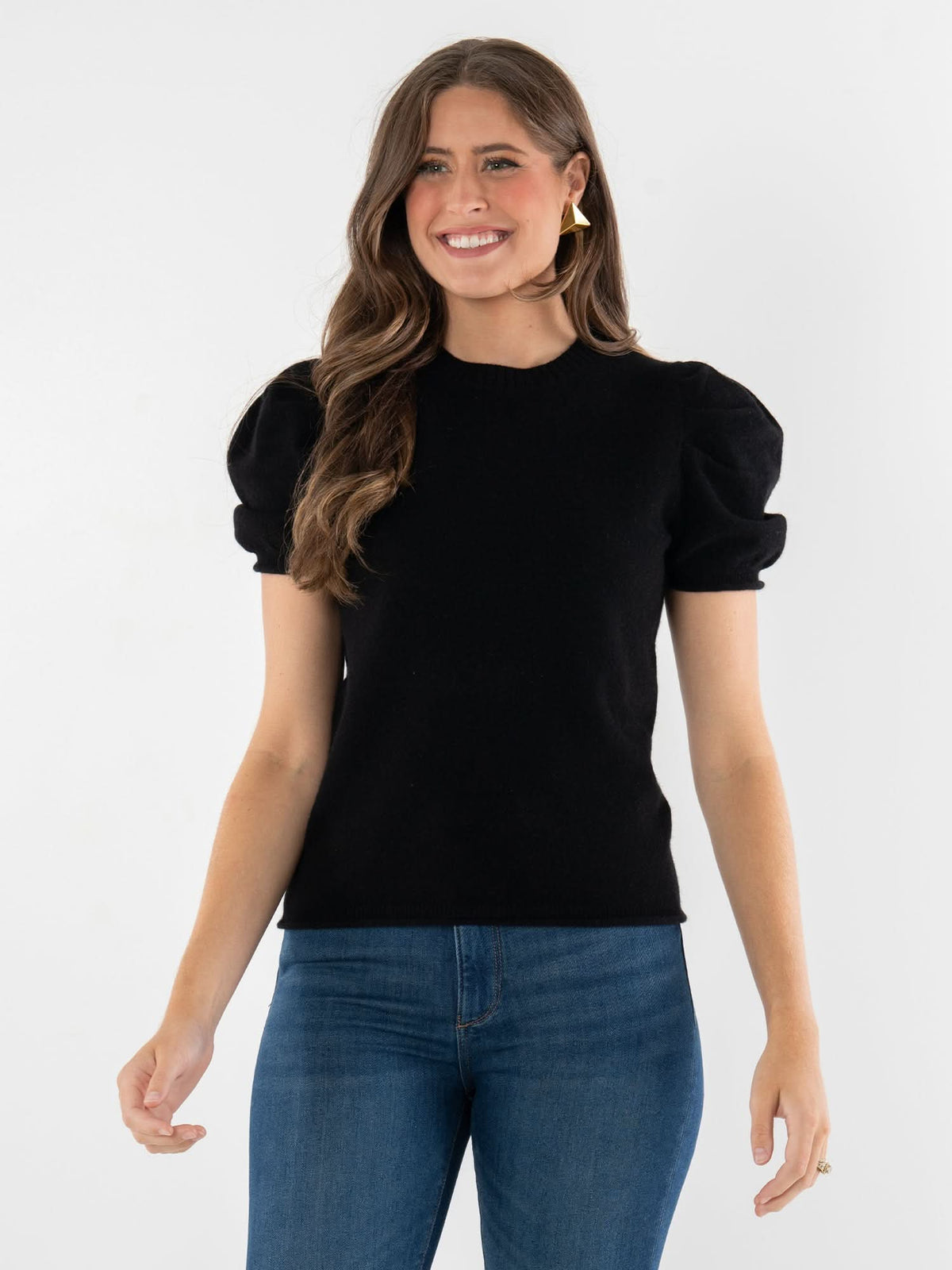 A woman with long brown hair is smiling, wearing a luxury Crewneck Tee with puff sleeves and blue jeans, standing against a plain white background.