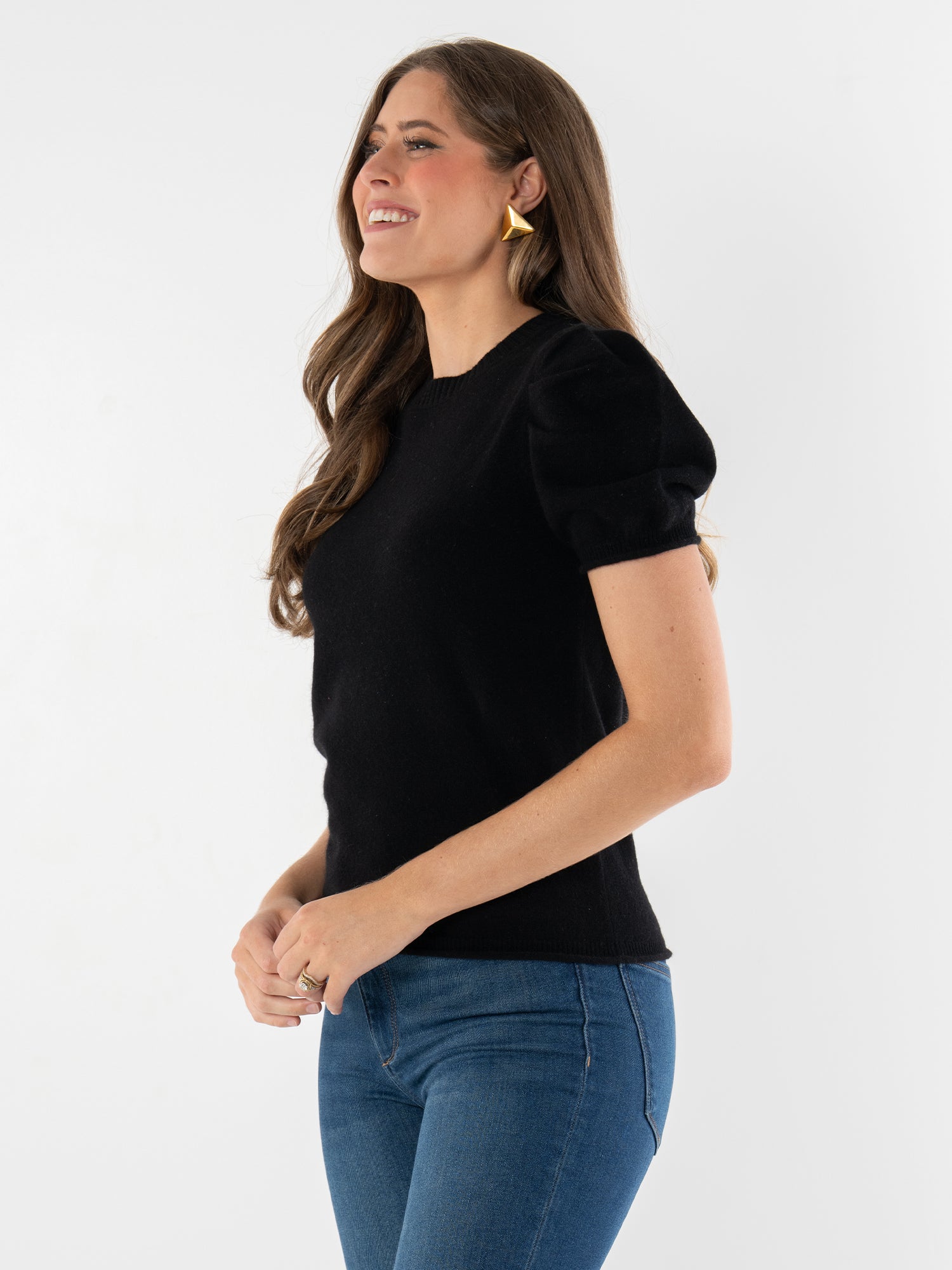 A woman with long brown hair wears the Crewneck Tee with puff sleeves and blue jeans, smiling against a plain white background. She accessorizes with gold geometric earrings and a ring on her finger.