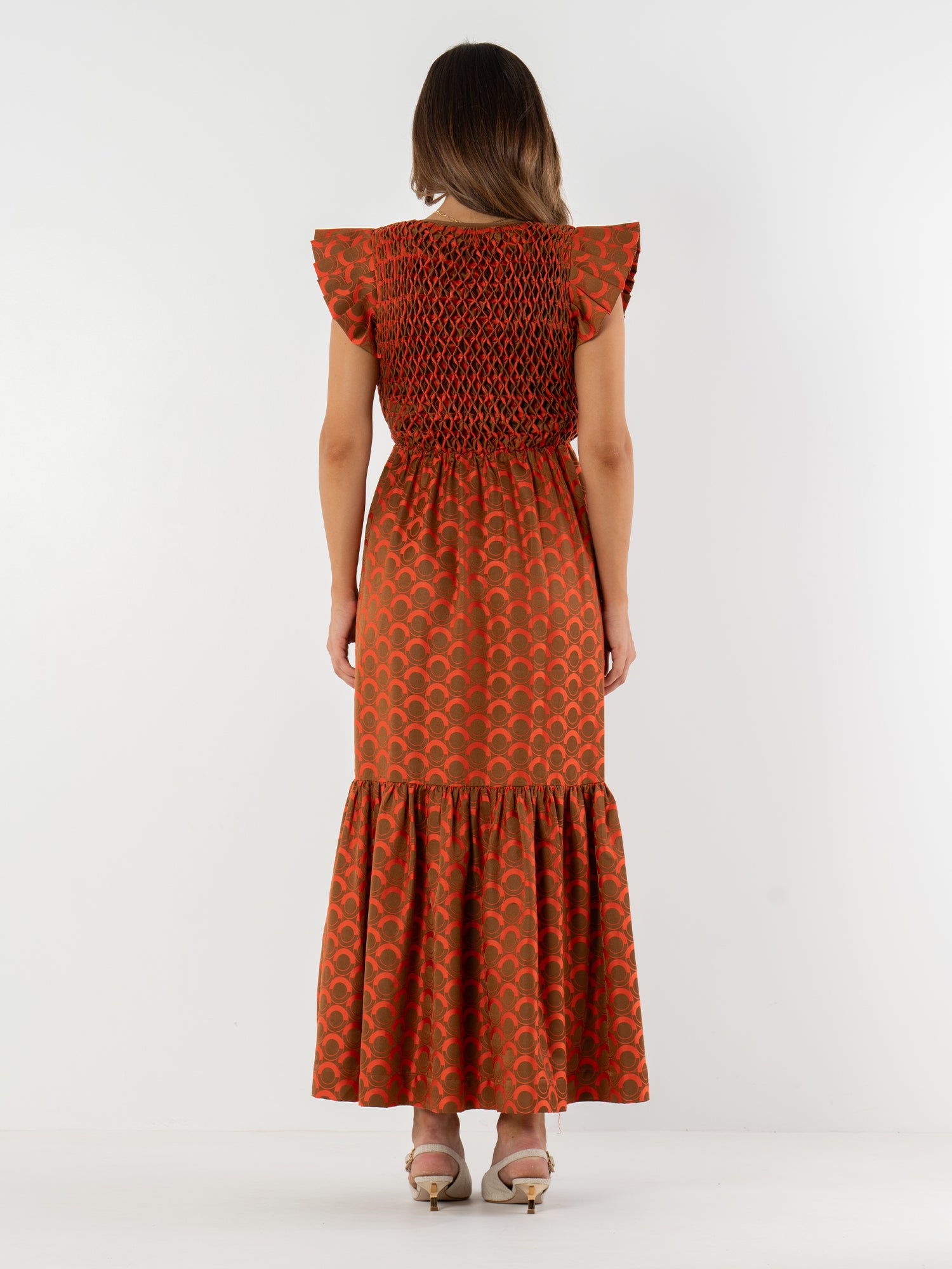 A woman faces away, wearing the vibrant orange Grace Maxi with a bold pattern, ruffled sleeves, smocked bodice, and flared hem—evoking vintage Italian style. She completes the look with heeled sandals.