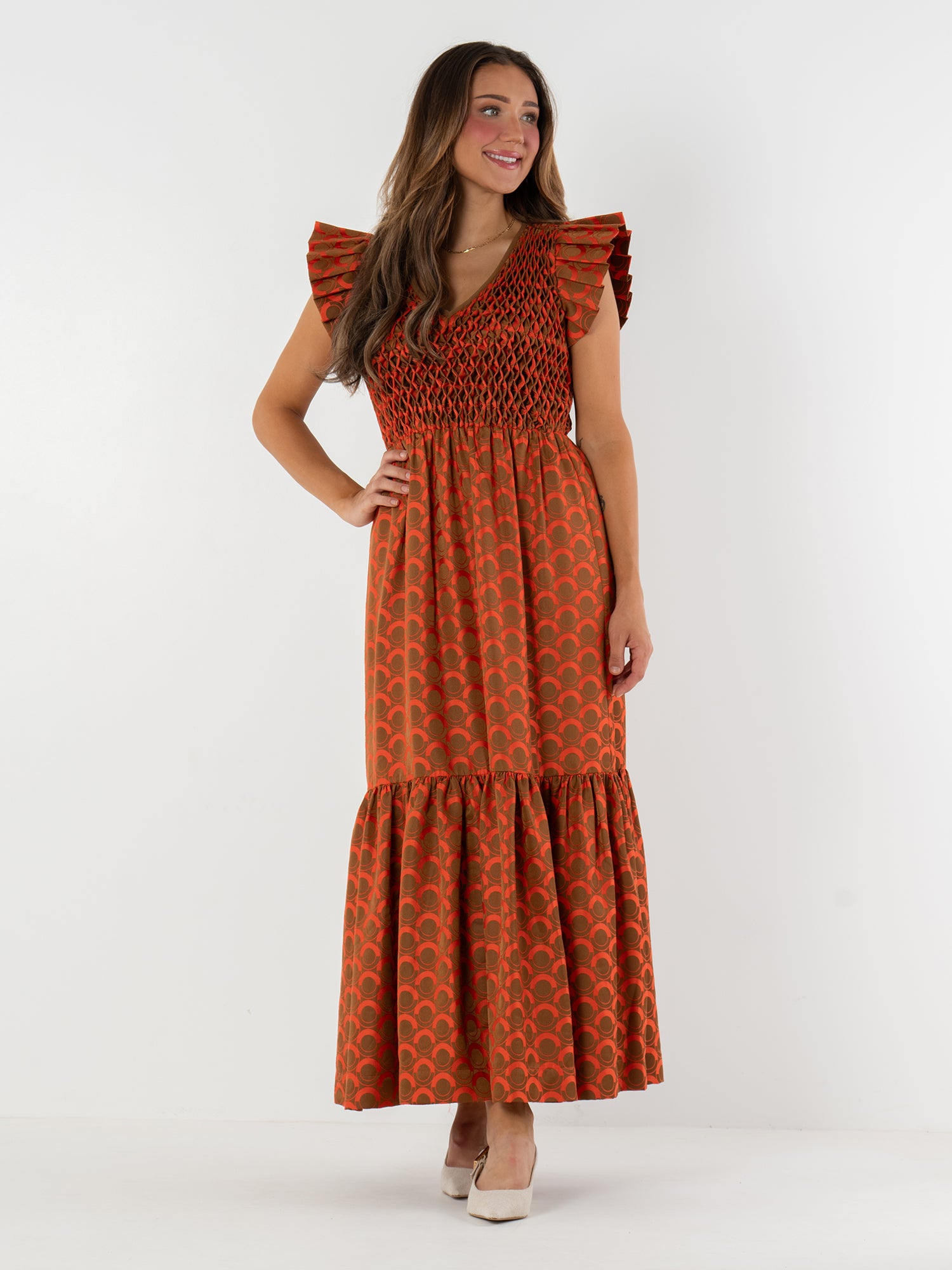 Against a white background, a woman wears the Grace Maxi—an orange maxi dress with a red scallop print, ruffled cap sleeves, and shirred bodice. She smiles in white heels as her long brown hair flows over the vintage-inspired style.