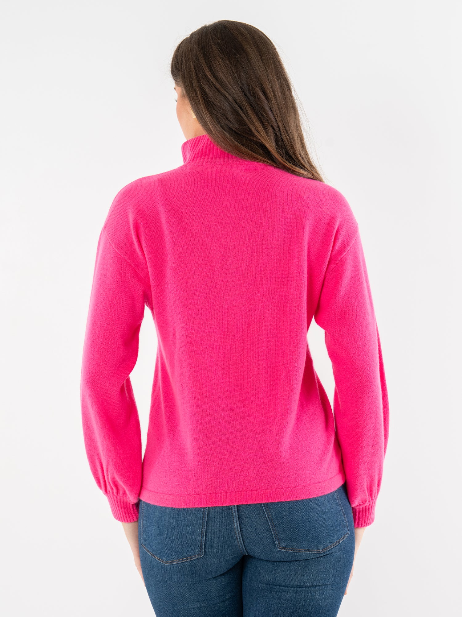 A woman with long brown hair, wearing the bright pink Lolli Sweater and blue jeans, stands with her back to the camera against a plain white background.