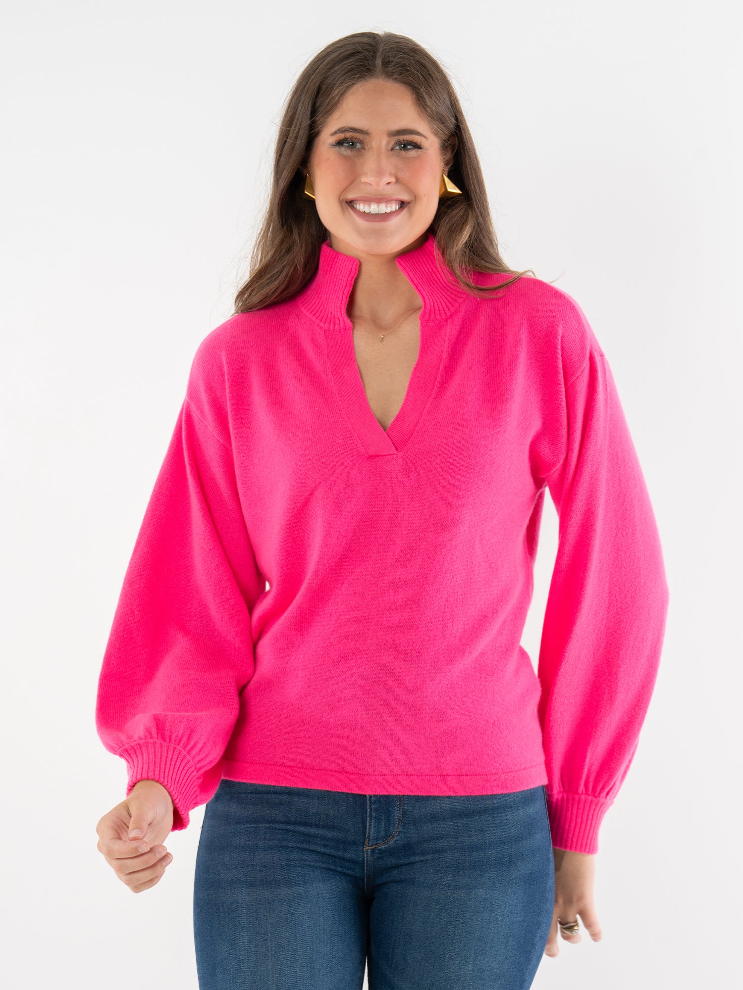A woman with long brown hair smiles while wearing the Lolli Sweater, a bright pink cashmere top featuring a split collar and balloon sleeves, paired with blue jeans against a plain white background.