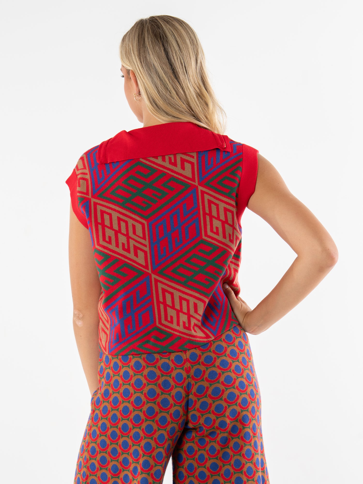 A woman with long blonde hair stands facing away, wearing the Poppy Pullover Vest—red, sleeveless, and featuring a bold geometric pattern in blue, red, and tan—paired with high-waisted blue and red patterned pants.