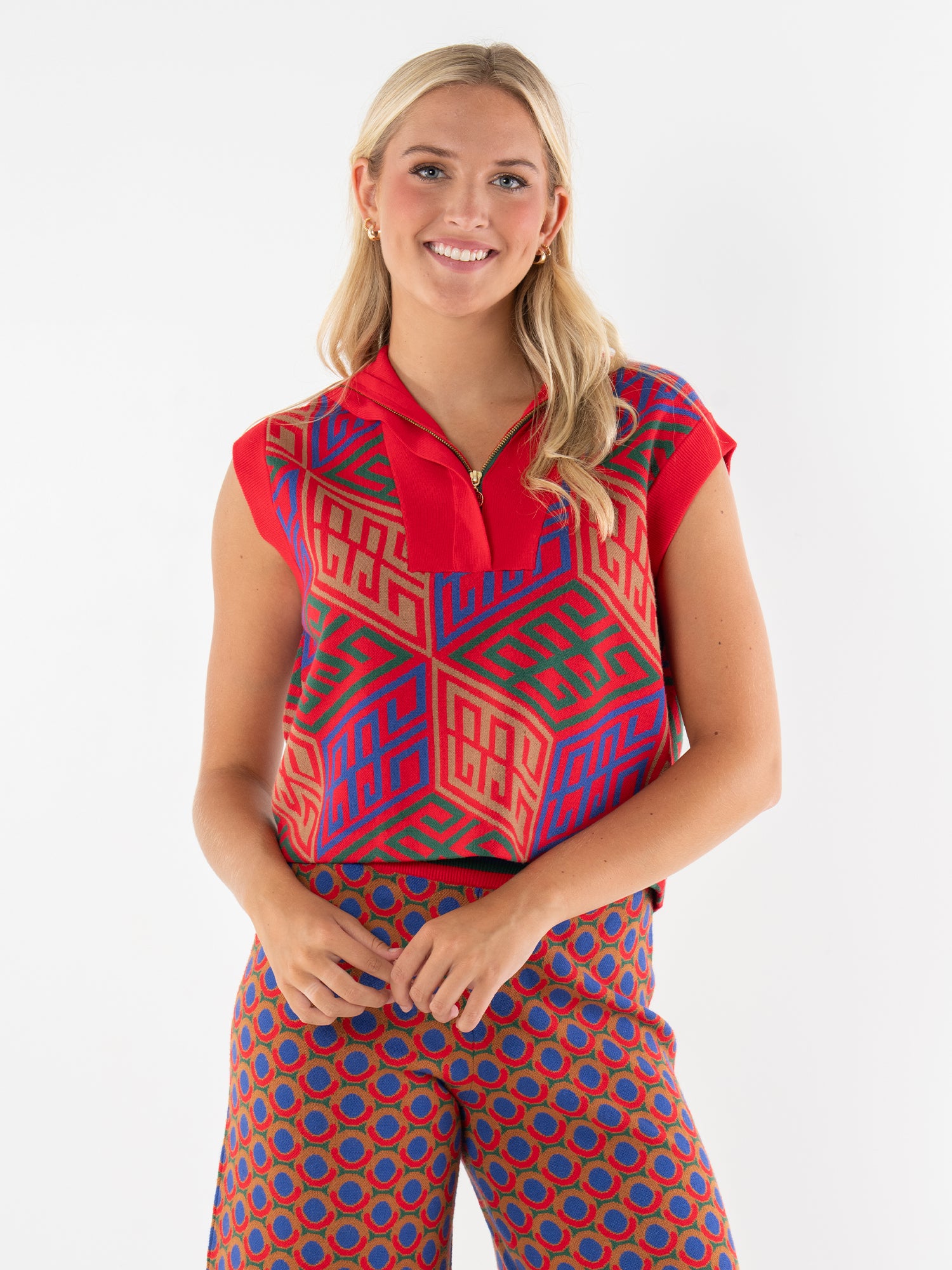 A smiling woman with long blonde hair wears the Poppy Pullover Vest, a colorful sleeveless top with red, blue, and green geometric patterns and an oversized collar, paired with matching red and blue patterned pants against a plain white background.