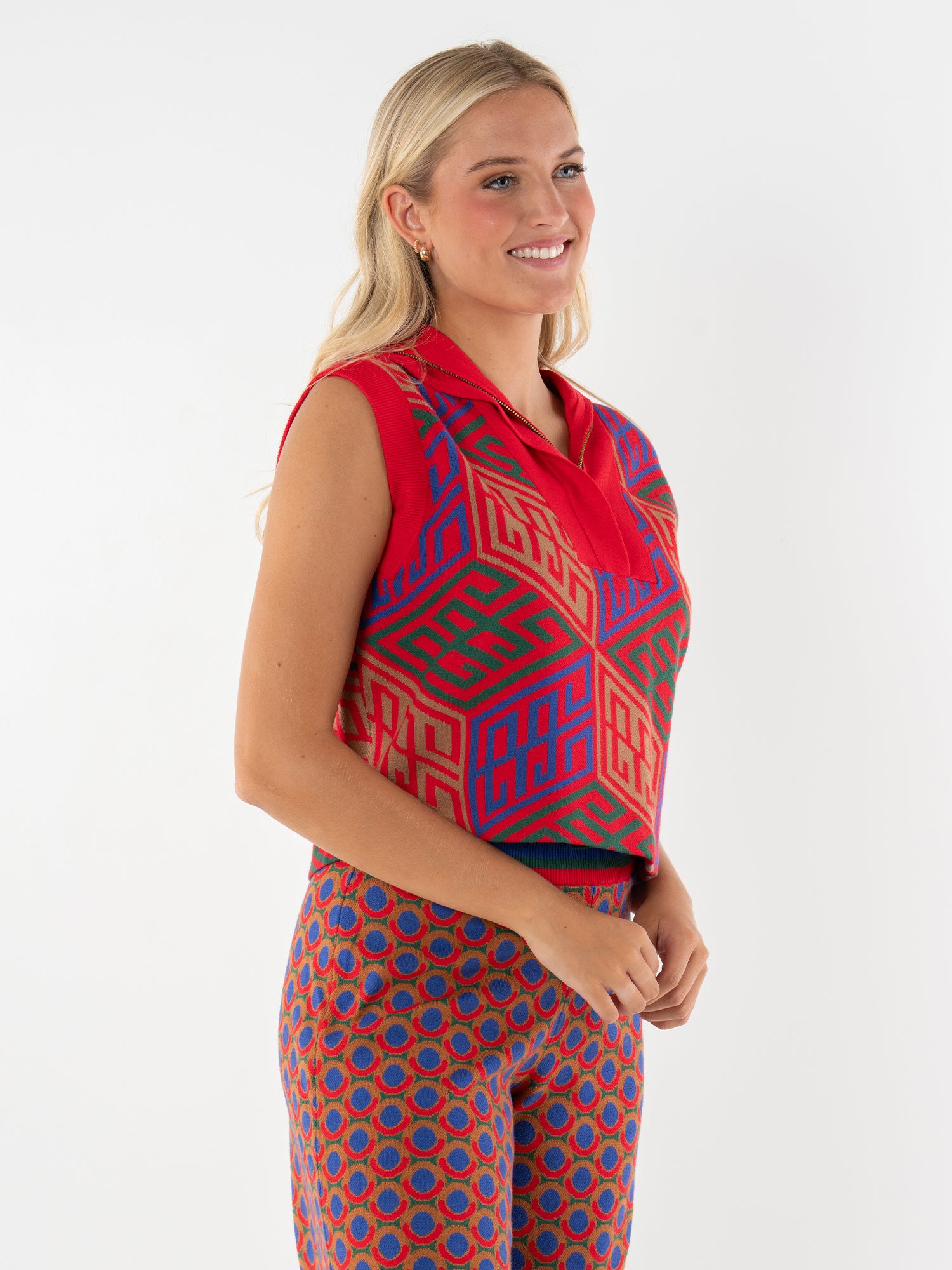 A woman with long blonde hair smiles while wearing the Poppy Pullover Vest—a sleeveless top with red, blue, and green geometric patterns and an oversized collar—against a plain white background.