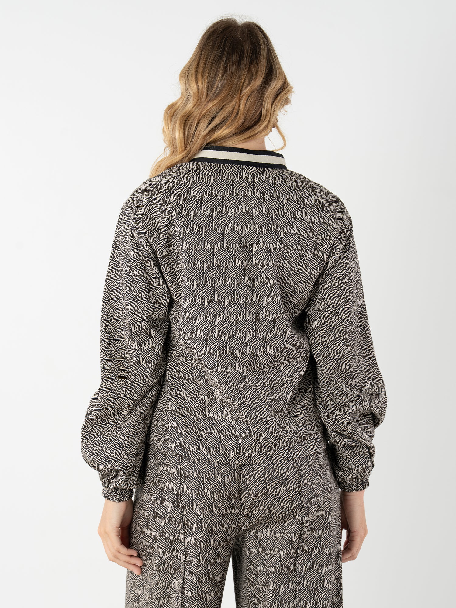 A woman with wavy blonde hair stands facing away, wearing the Lolli Top—a patterned long-sleeve piece with relaxed dolman sleeves and a black and white collar—against a plain white background.