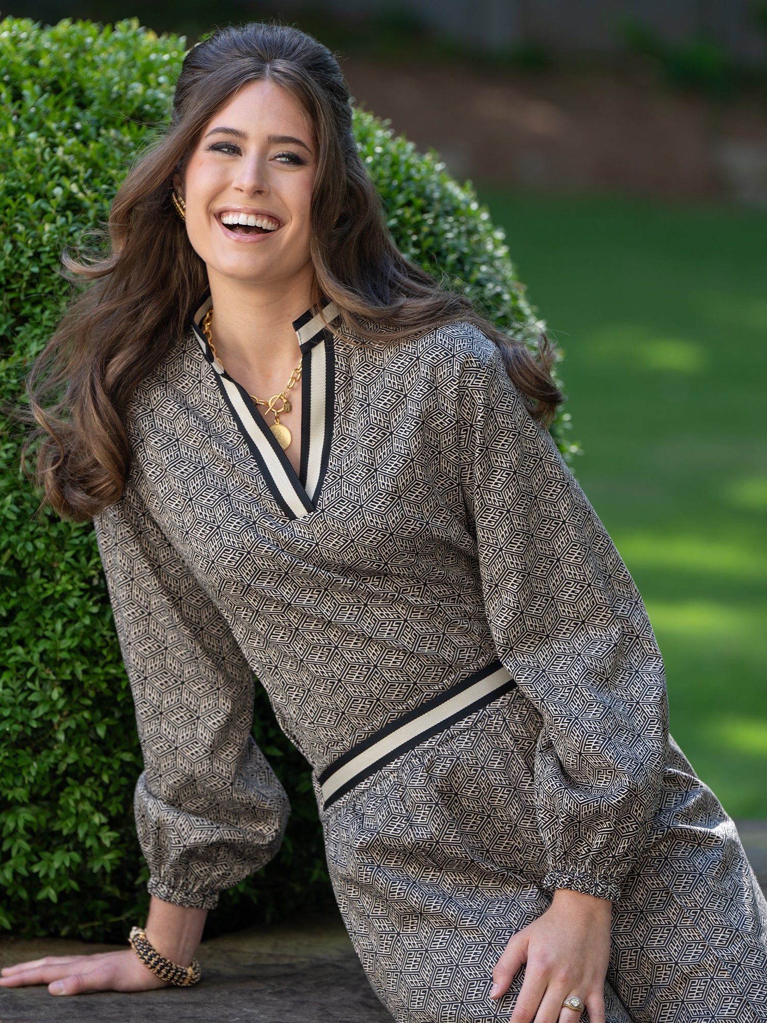 A woman with long brown hair wears the Lolli Top with relaxed dolman sleeves, leaning against a stone surface and smiling outdoors, with green shrubs and grass in the background.