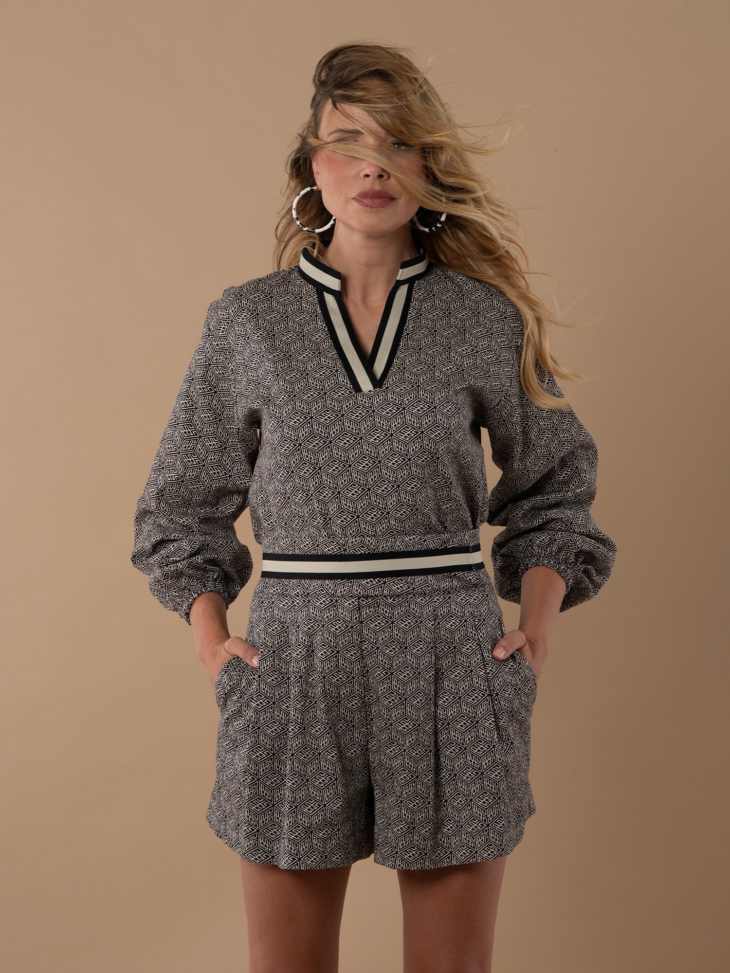 A woman stands against a beige background wearing the Lolli Top, a patterned long-sleeve piece with relaxed dolman sleeves, black and white collar and waistband. Her hands are in her pockets as her hair blows across her face.
