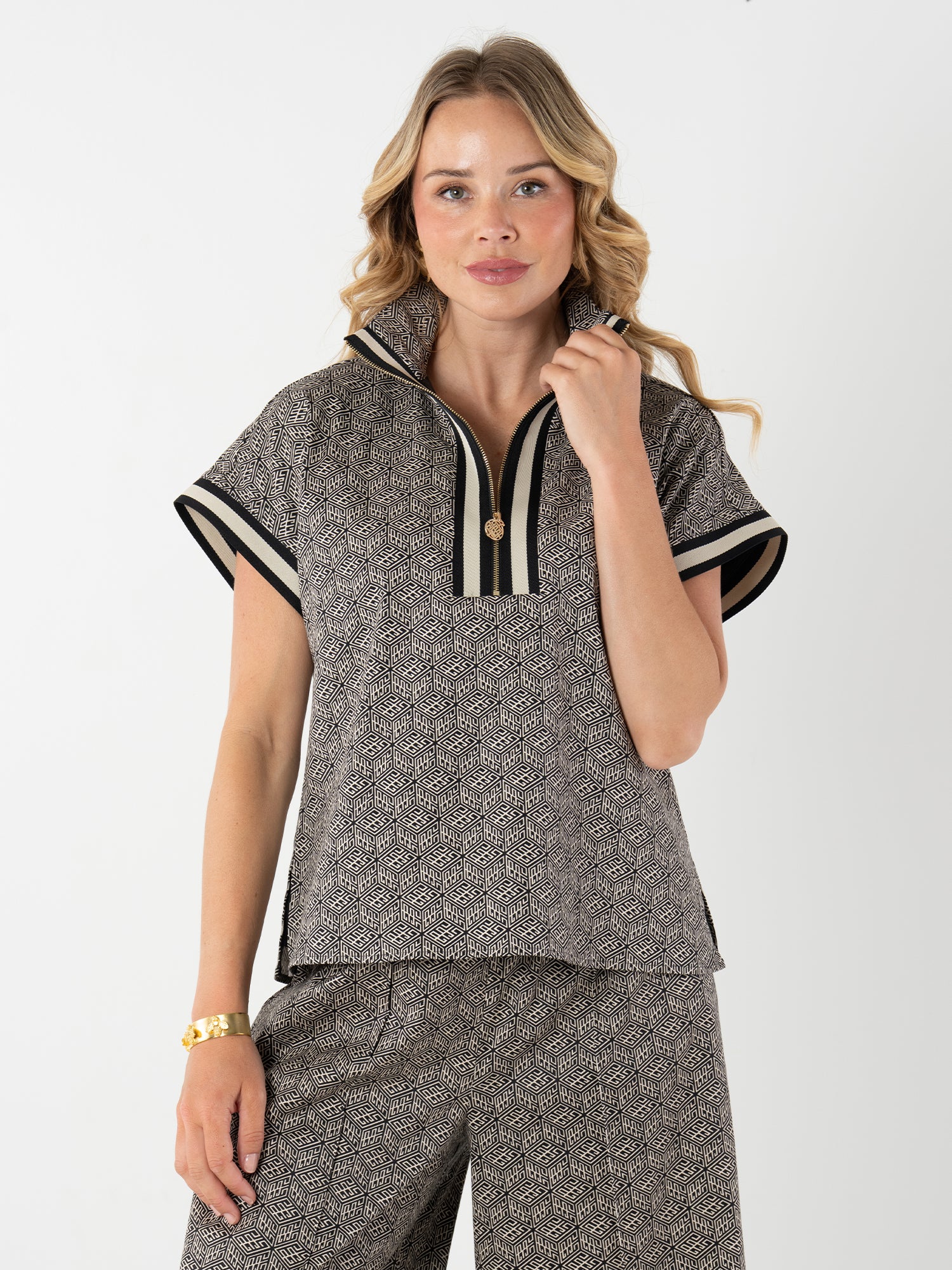 A woman with wavy blonde hair models the Poppy Pullover—a women’s patterned pullover with a striped collar—paired with coordinating wide-leg pants, posing confidently in front of a plain white background.