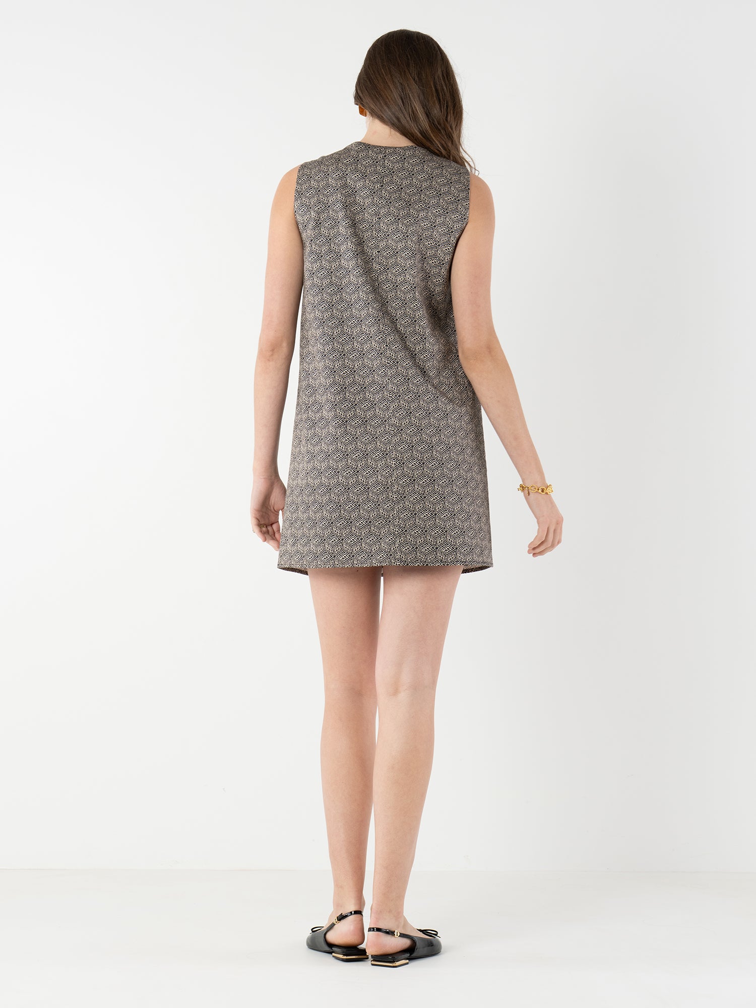 A woman with long brown hair, wearing the Tory Dress—a sleeveless, patterned shift—paired with black sandals and a gold bracelet, poses against a plain white background.