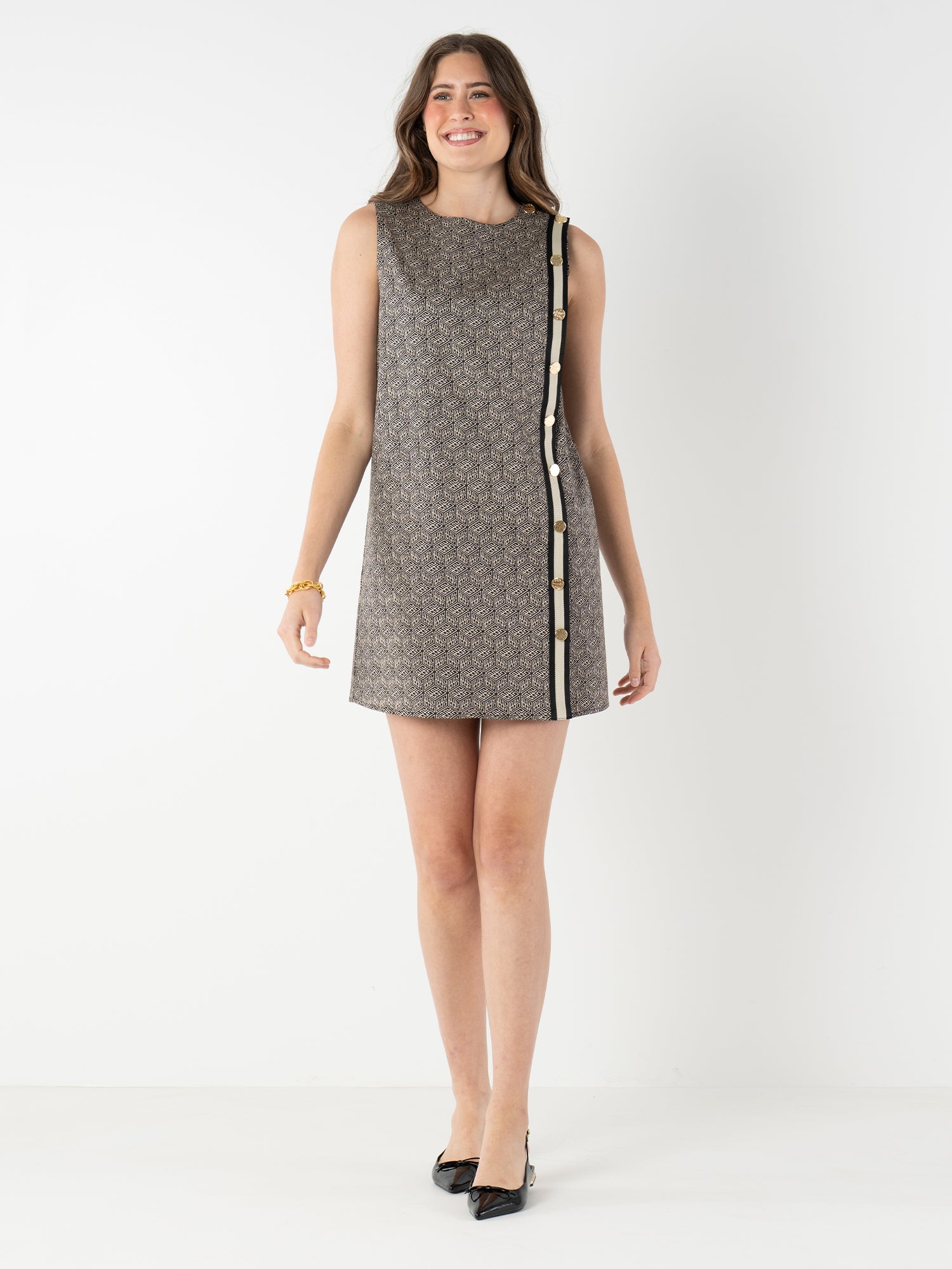 A woman stands against a white background, smiling and wearing the Tory Dress—a sleeveless, patterned shift dress with side button details—paired with black pointed-toe heels.