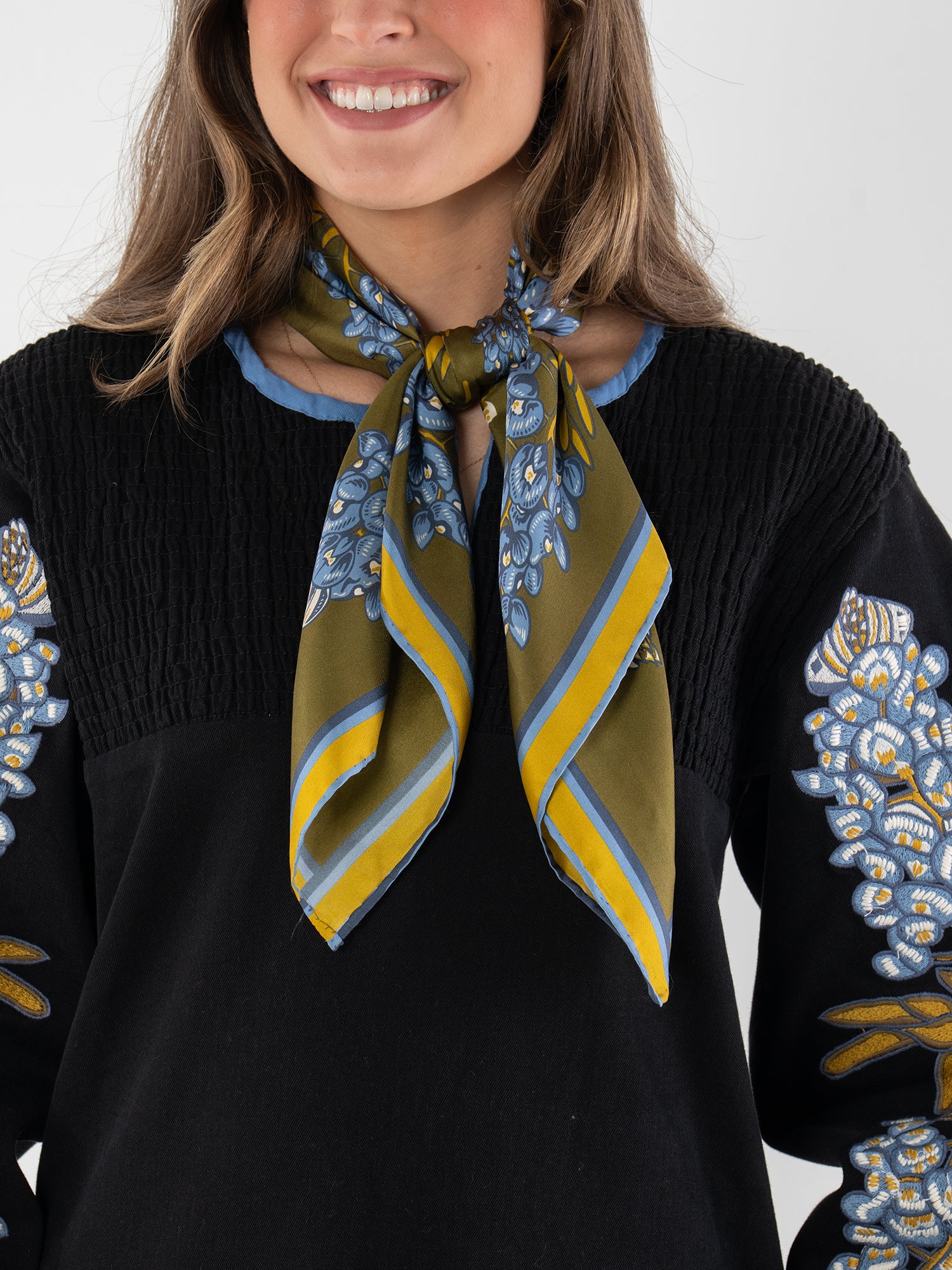 A woman with wavy brown hair smiles, wearing a black top with blue and white floral embroidery and a Hand-Stitched Silk Scarf in green silk with blue and yellow stripes—an elegant nod to the Texas state flower—tied around her neck.