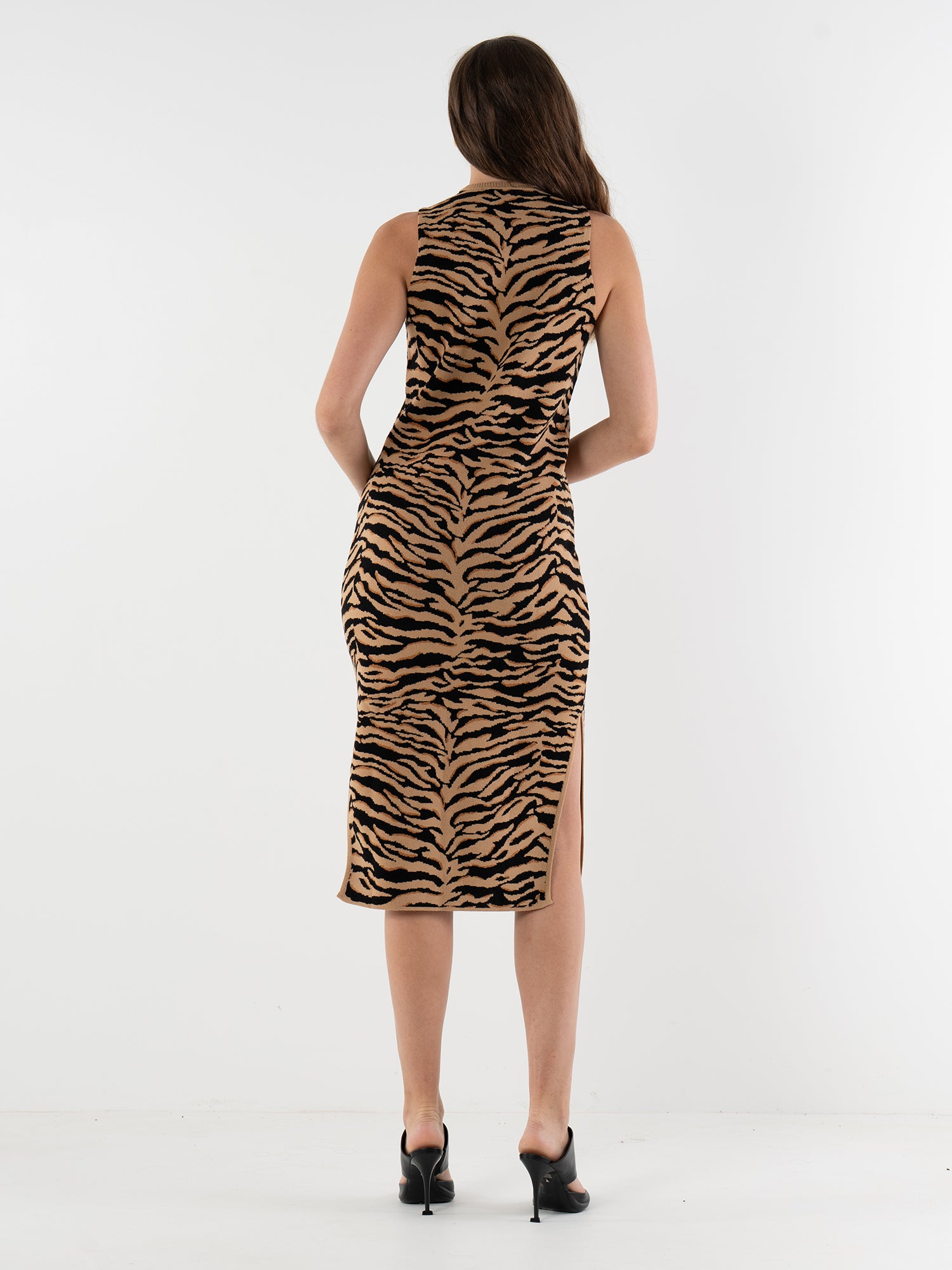 A woman with long brown hair, wearing the Simone Sweater Dress—a sleeveless, knee-length tiger print dress with a side slit—stands with her back to the camera in black heeled mules against a white background.