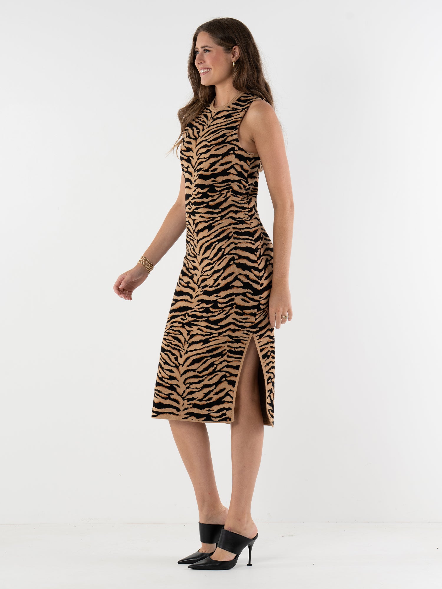 A woman with long brown hair and black heeled mules stands sideways, smiling, in the Simone Sweater Dress—a sleeveless, knee-length tiger print viscose knit dress with a side slit—against a plain white background.