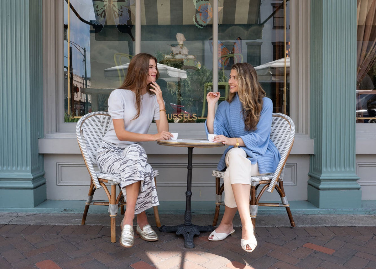 Two women sitting at a small outdoor cafe table in front of a storefront with a striped awning, brick sidewalk, and large windows showing butterfly artwork.