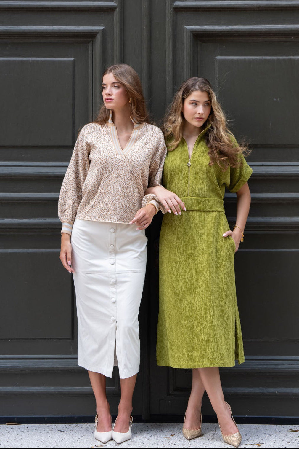 Two women standing in front of a large dark green paneled door. The woman on the left wears a beige knit long-sleeve top and a white button-front midi skirt.