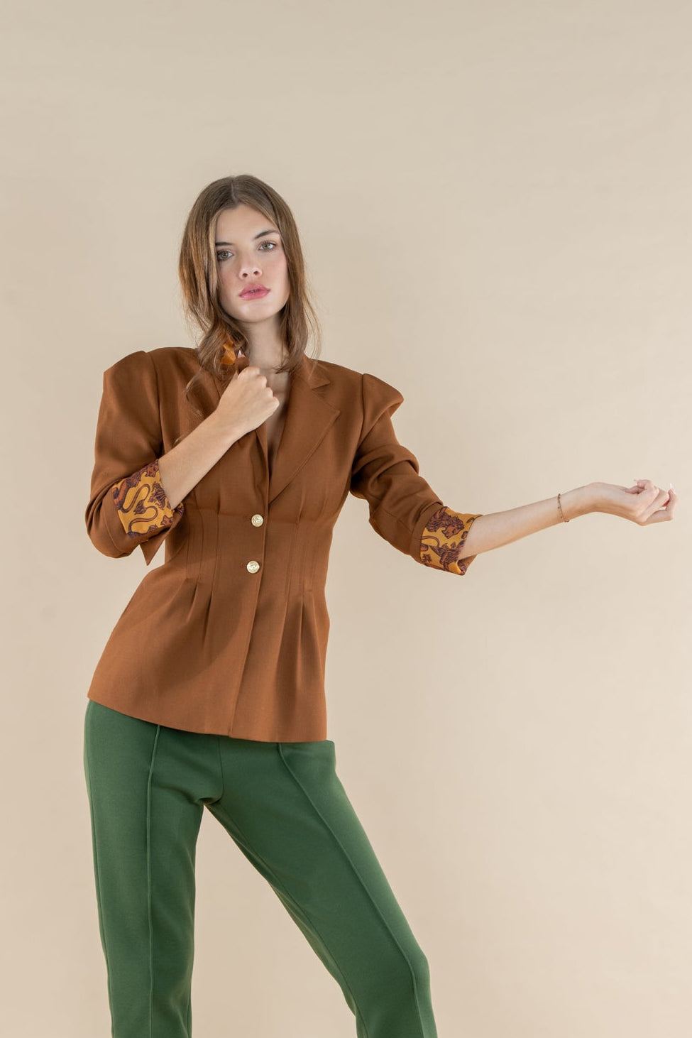 A woman modeling a brown cropped blazer with gold buttons and patterned sleeves, paired with green cropped trousers and beige heels, against a solid beige studio background.