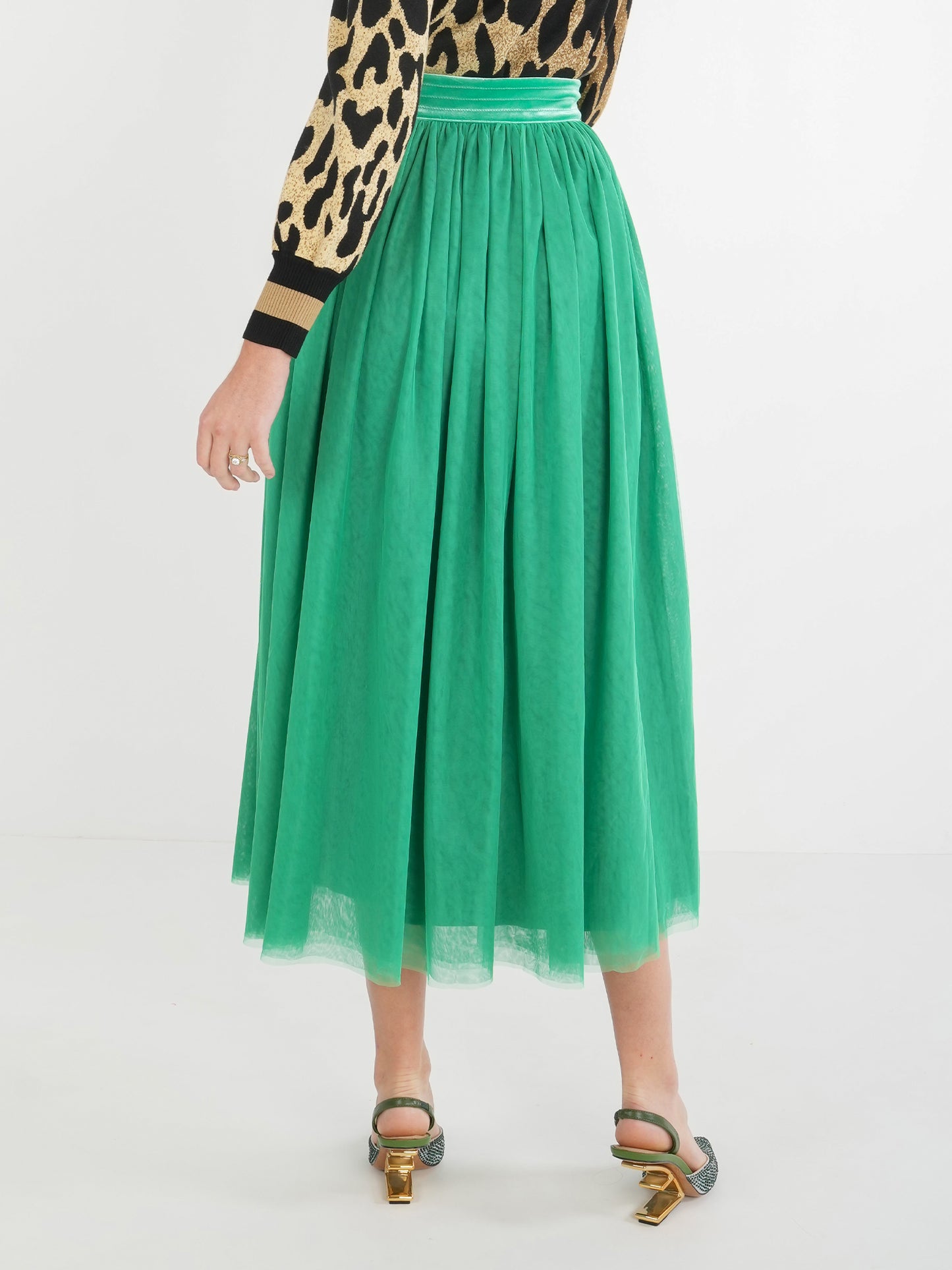 A person wearing a leopard print top and a long, green pleated Misty Maxi - Jolly Green stands on a white background. The skirt, accentuated by a velvet waistband, is paired with open-toed heels. Only the back of the person is visible.