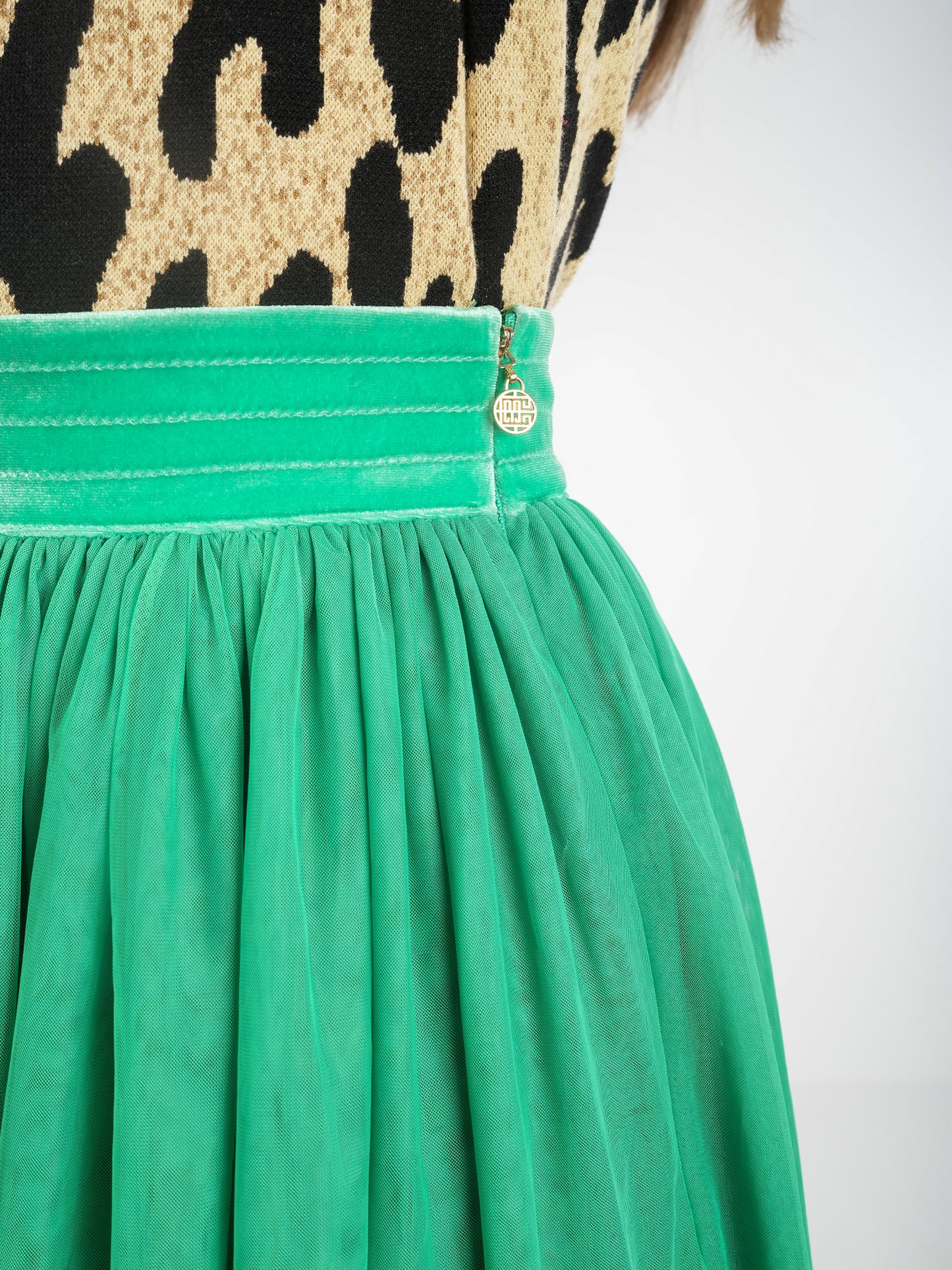 Close-up of a person wearing a leopard print top paired with the Misty Maxi in Jolly Green, featuring a green pleated skirt with a velvet waistband. A small circular pendant near the waist enhances the charm of the ensemble against a plain white background.