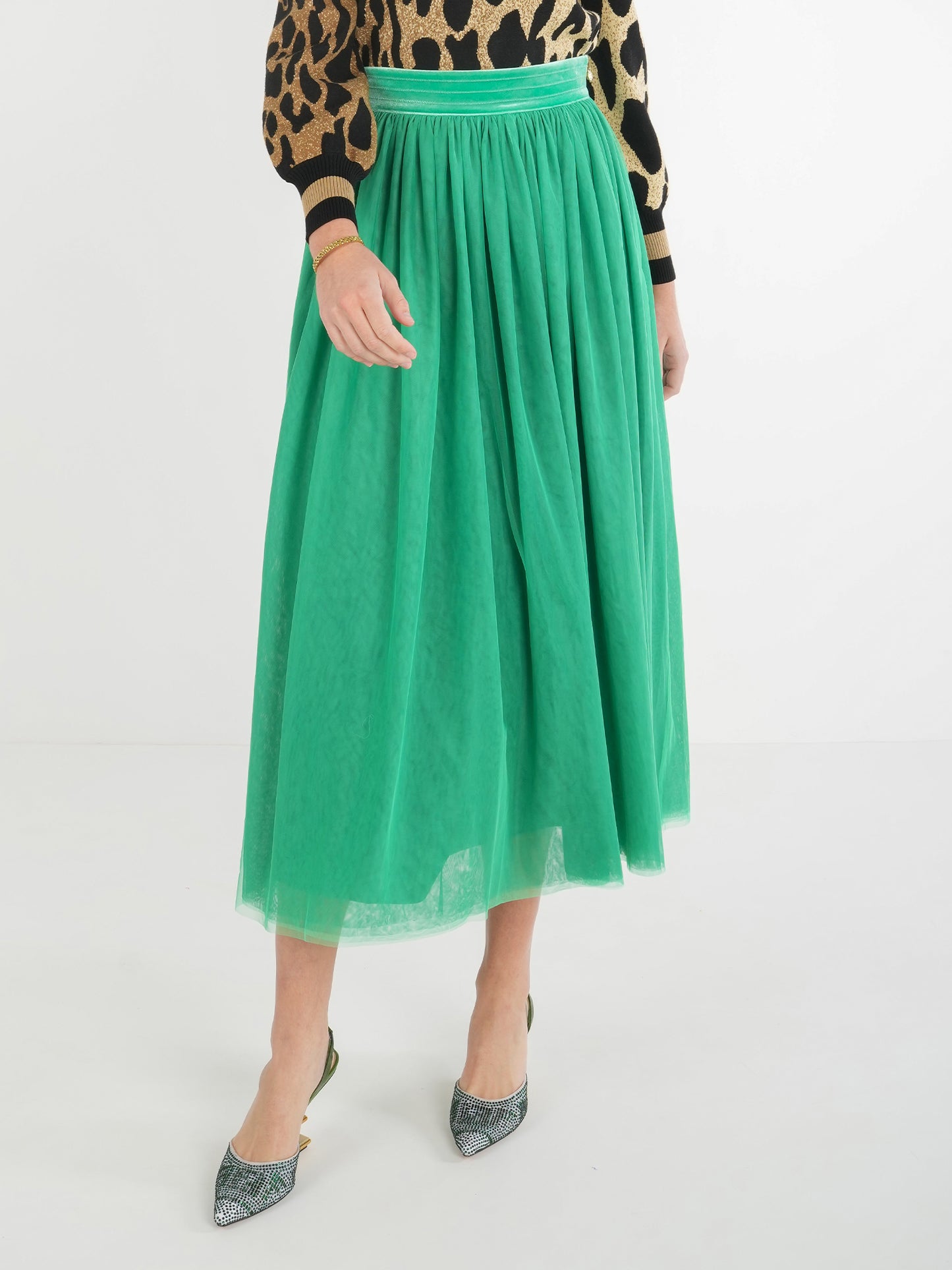 A person is wearing the Misty Maxi - Jolly Green, a midi skirt with a green tulle design and a velvet waistband, paired with a leopard print sweater and pointed shoes featuring a metallic pattern, standing against a plain white background.