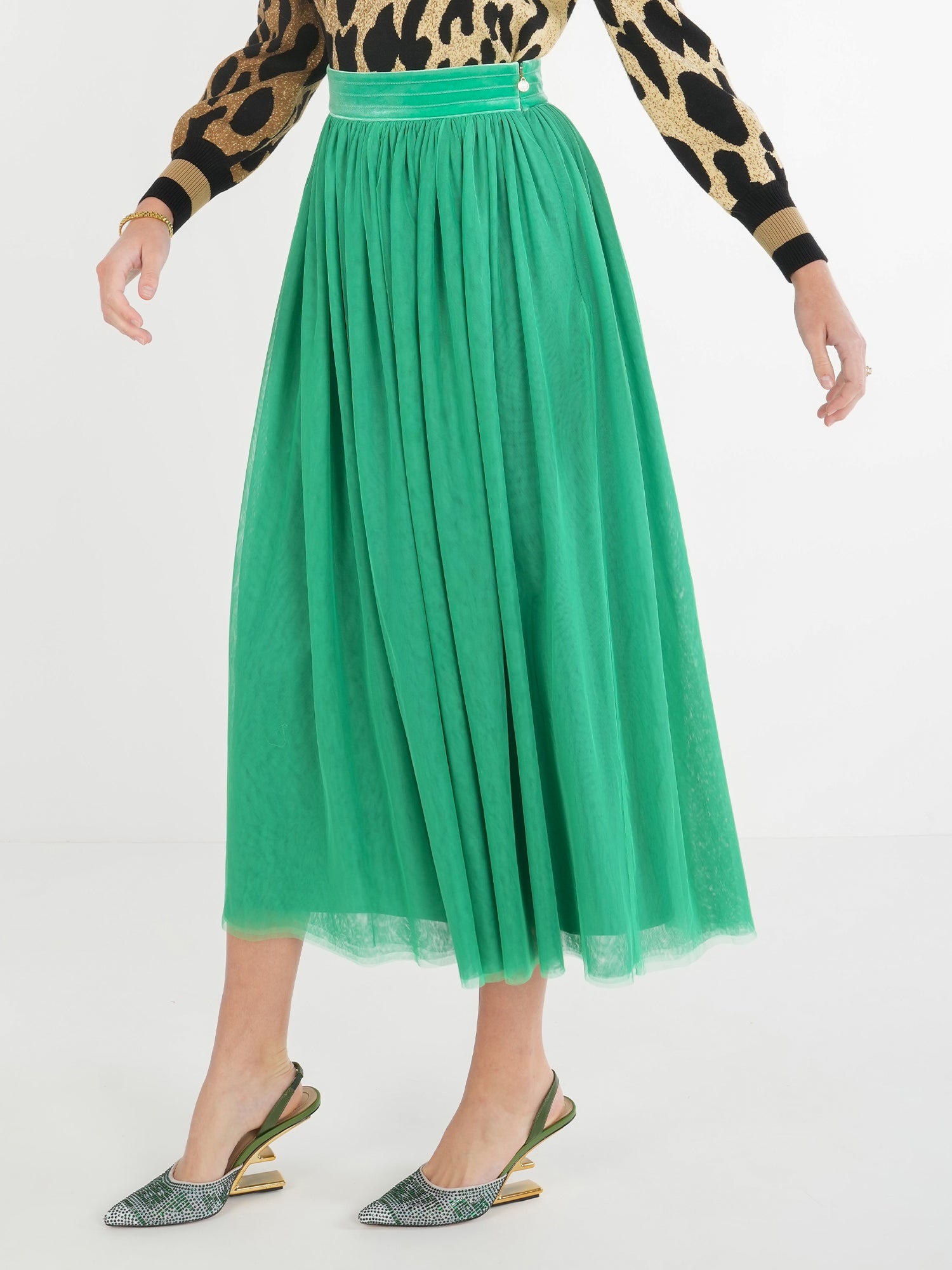 A person is wearing the Misty Maxi - Jolly Green skirt paired with a leopard print sweater and a velvet waistband. The skirt flows gracefully, and they have completed the outfit with metallic-finished pointed heels for an elegant touch. The background features a plain white wall.