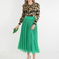 A woman in a leopard-print sweater and a Misty Maxi - Jolly Green Skirt featuring a velvet waistband holds a small green clutch. She completes her ensemble with green high heels and a matching headband, all while standing gracefully against a white background.