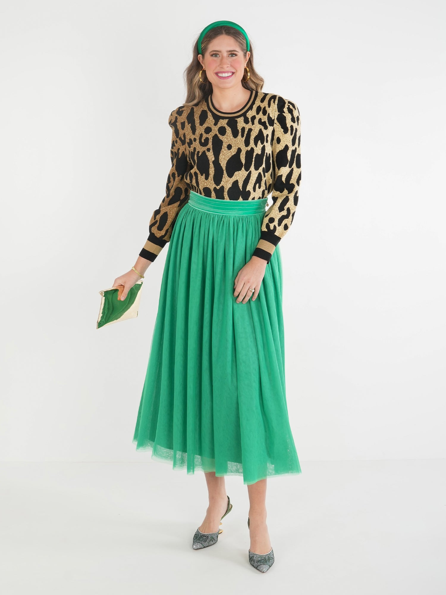 A woman in a leopard-print sweater and a Misty Maxi - Jolly Green Skirt featuring a velvet waistband holds a small green clutch. She completes her ensemble with green high heels and a matching headband, all while standing gracefully against a white background.