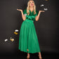 A woman wearing a shiny green blouse and the Misty Maxi - Jolly Green skirt, featuring a velvet waistband, smiles while raising her hands. Gold and silver metallic bows float around her against a black background, adding a touch of magic. She completes the look with elegant gold heels.