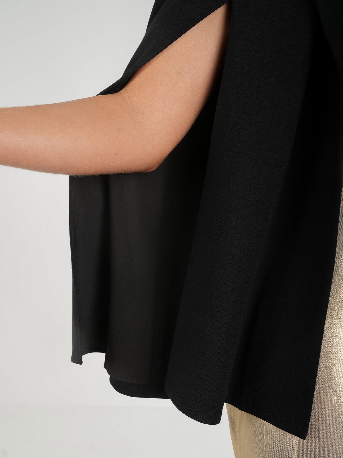 Close-up of a person wearing the Overlay Cape - Silent Night, a sleeveless black top with a flowing cape design at the shoulder, paired with metallic gold pants. The image focuses on the side view of the garment and the model's arm against a neutral background.