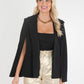 A blonde woman radiates elegance, smiling as she dons the Silent Night Overlay Cape over her square-neck top and a black blazer. Her ensemble is completed by high-waisted metallic gold pants and a gold chain necklace, all set against a plain white background, reminiscent of Silent Night's serene charm.