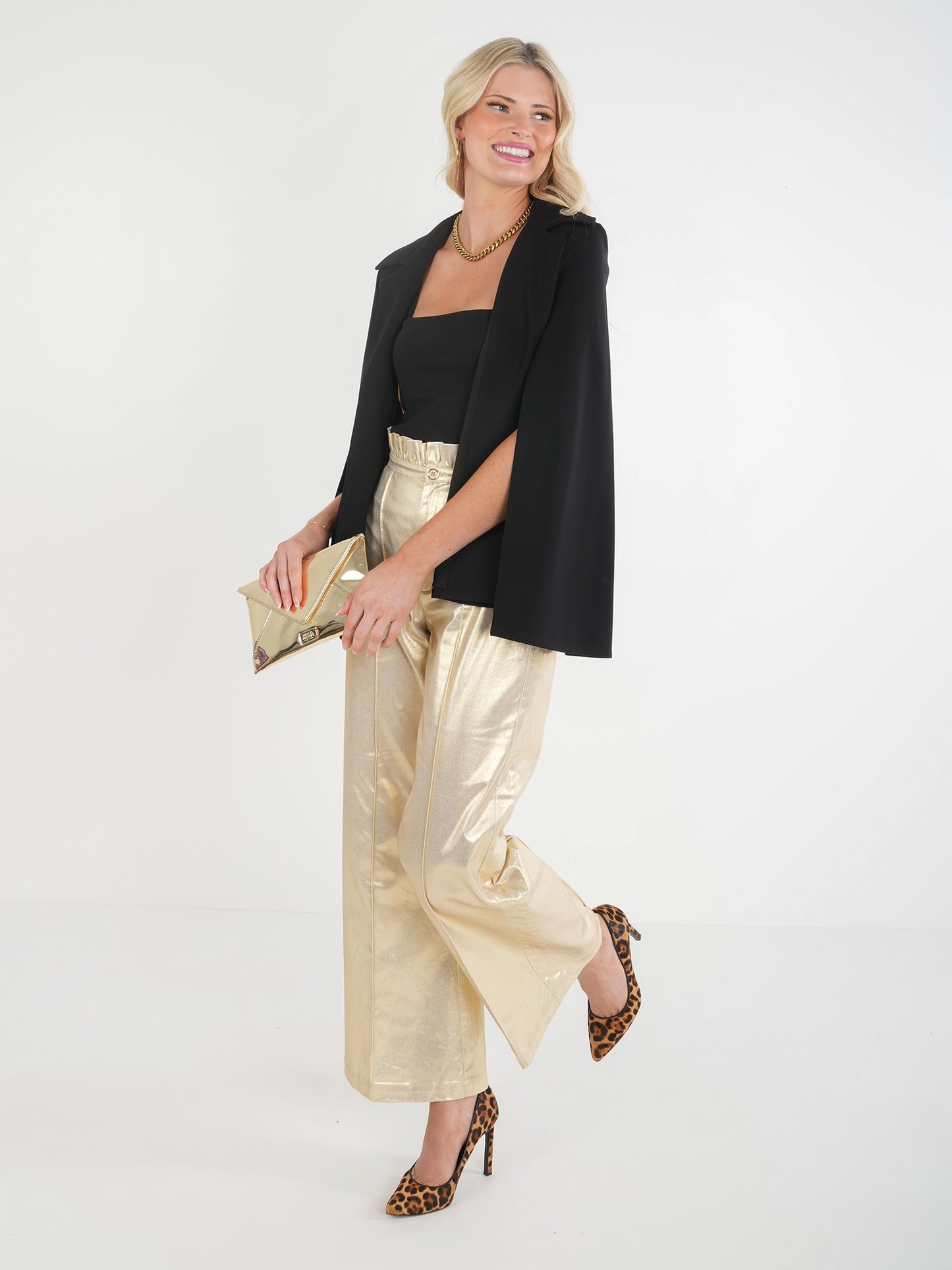 A person stands confidently, smiling, wearing the Overlay Cape - Silent Night featuring an elegant classic lapel over a black top and shiny gold pants. They hold a gold clutch and wear leopard-patterned high heels. Their hair is styled in loose waves.