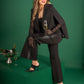 A woman in a stylish outfit featuring the Overlay Cape - Silent Night poses confidently with one leg up on a green block. She holds a champagne flute, while a bucket of champagne sits beside her. The solid green background complements her chic ensemble.