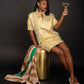 A woman in a gold-patterned Poppy Dress - Jaguar Jacquard sits on a matching stool against a black background. She holds a cocktail glass up in one hand, wearing white shoes, while a luxurious fur coat drapes over the stool.