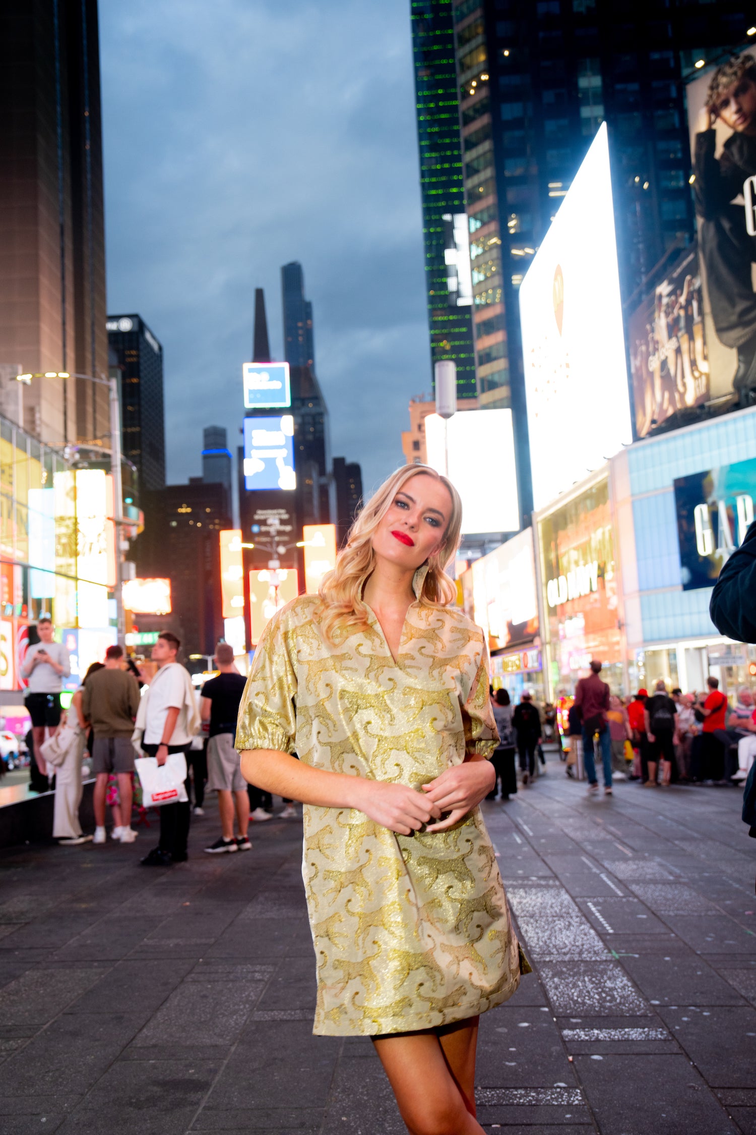 A woman in a stunning Poppy Dress - Jaguar Jacquard poses confidently in a bustling Times Square at dusk, surrounded by bright billboard lights and people.