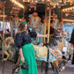 A woman in a black blouse and the Jolly Green Misty Maxi skirt, featuring a velvet waistband, poses beside a vintage carousel horse. She holds a handbag as colorful lights illuminate the festive scene. Other intricate carousel decorations are visible in the background, enhancing the magical atmosphere.