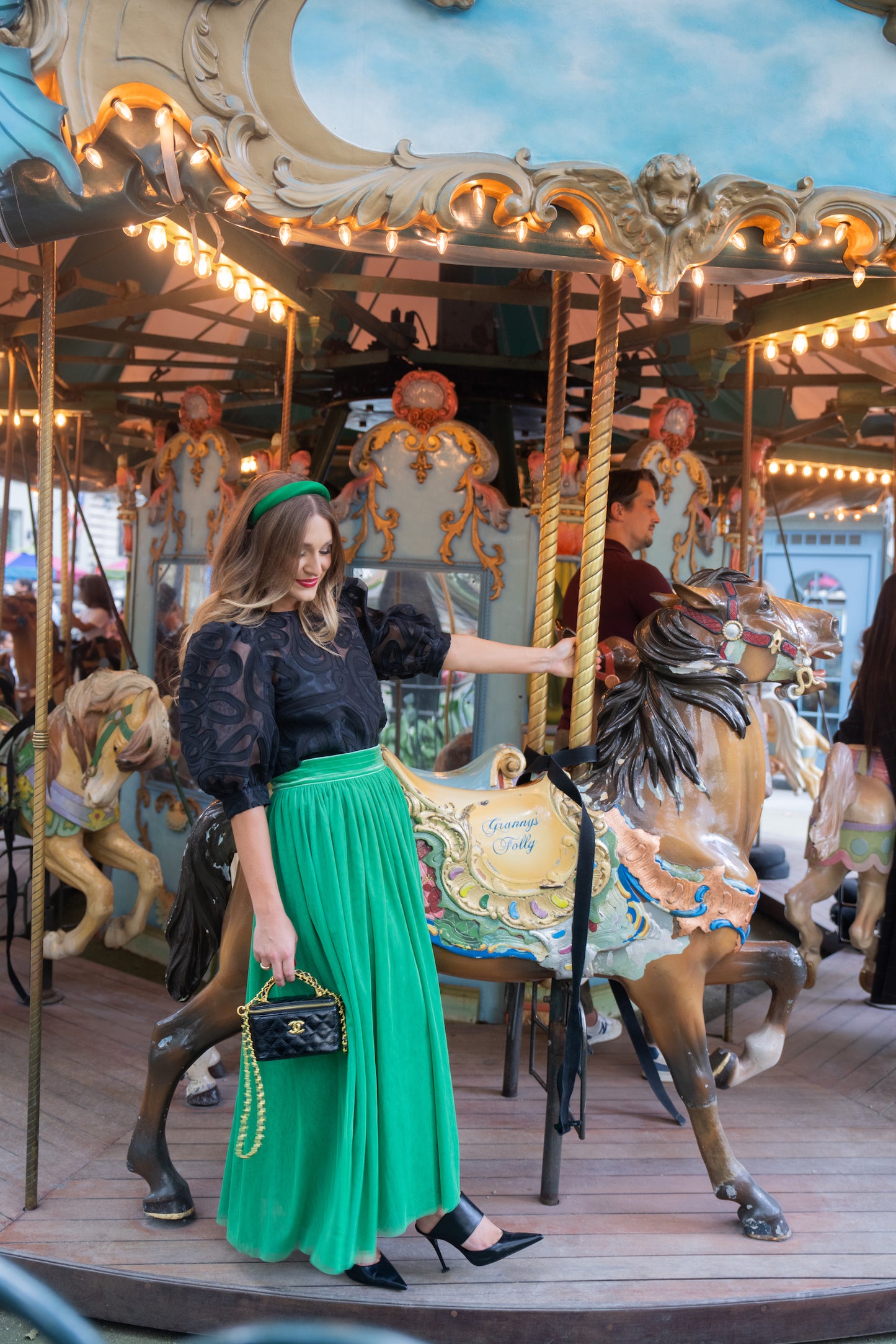 A woman in a black blouse and the Jolly Green Misty Maxi skirt, featuring a velvet waistband, poses beside a vintage carousel horse. She holds a handbag as colorful lights illuminate the festive scene. Other intricate carousel decorations are visible in the background, enhancing the magical atmosphere.