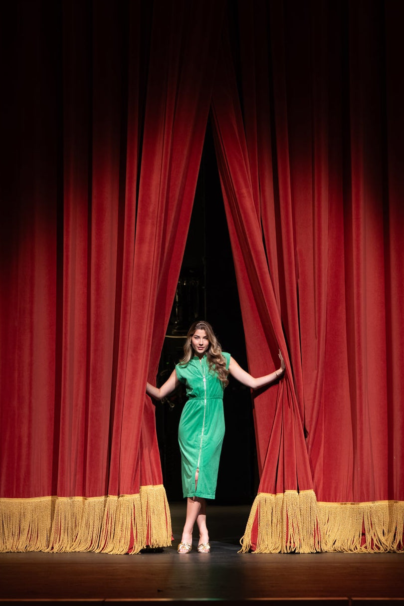 A woman in a bright green dress stands on a stage opening a red curtain, with a spotlight illuminating her against a dark auditorium background.
