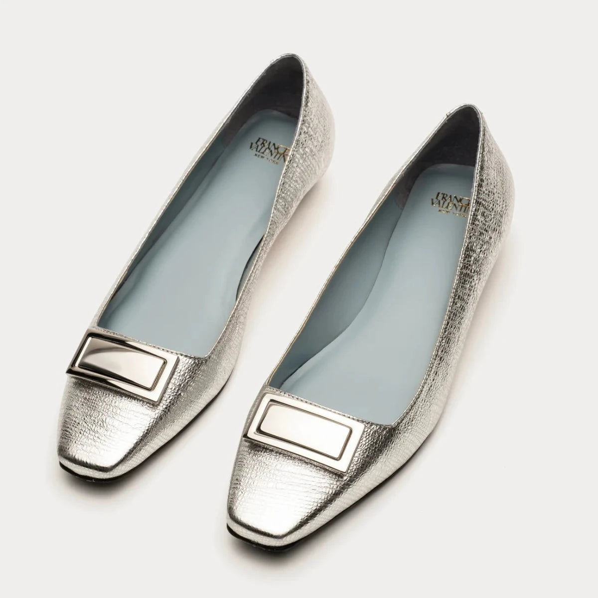 The Midge Flat Crinkled Patent Leather - Silver shoes showcase a stylish square-toe design and feature rectangular decorative buckles. Made from crinkle patent leather, they are beautifully presented on a light gray background.