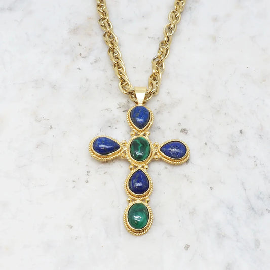 The Faith Turquoise Necklace by Niiki Paris is a gold chain necklace featuring a cross pendant adorned with blue and green gemstones. Its ornate design includes pear-shaped blue stones at the tips and a central green stone, all set against a light marbled background.