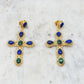 Faith Earrings, adorned with blue and green gemstones, are showcased on a white marble surface. These earrings draw inspiration from Parisian elegance, with each cross featuring a blend of teardrop and oval-shaped stones intricately set in detailed gold settings, capturing the chic allure of Niiki designs.