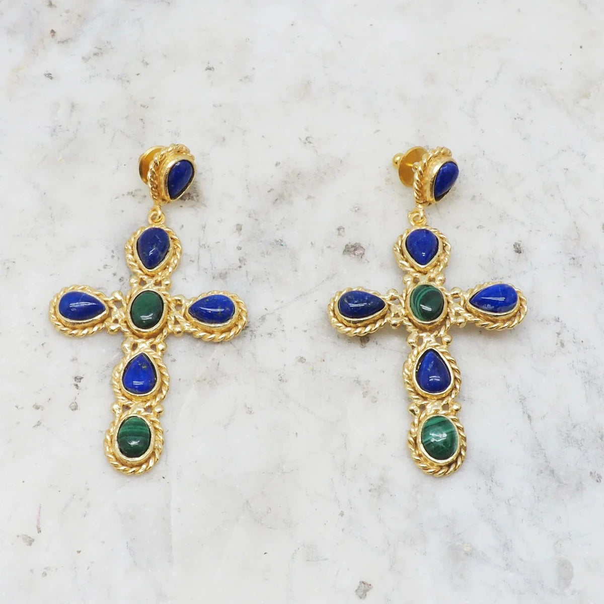 Faith Earrings, adorned with blue and green gemstones, are showcased on a white marble surface. These earrings draw inspiration from Parisian elegance, with each cross featuring a blend of teardrop and oval-shaped stones intricately set in detailed gold settings, capturing the chic allure of Niiki designs.