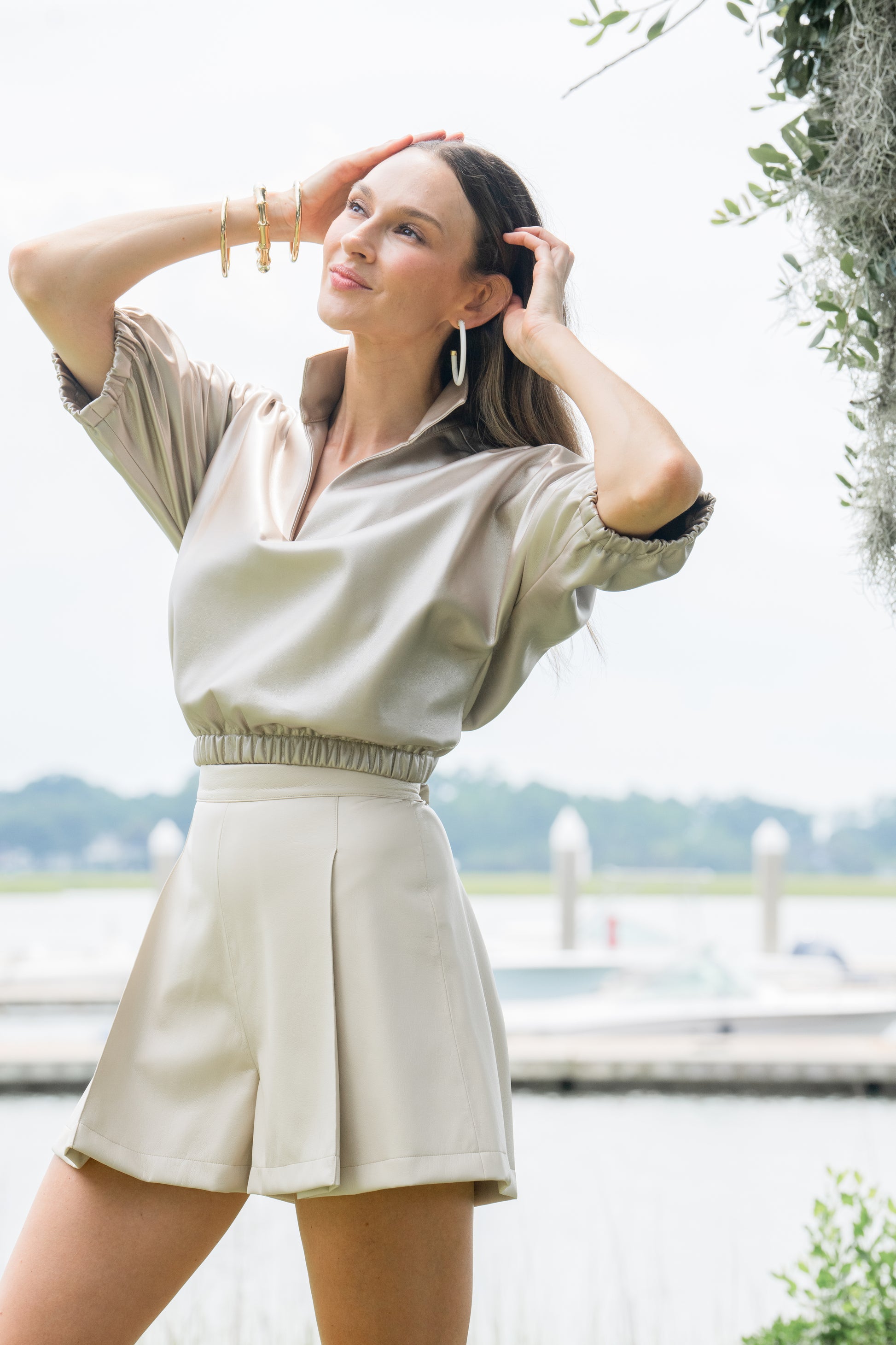 A woman in a light beige outfit stands outdoors near a waterfront, her cropped silhouette highlighting the shiny Poppy Crop - Pearl along with high-waisted shorts. She accessorizes with hoop earrings and bracelets, smiling amid the greenery and blurred backdrop of water and docks.