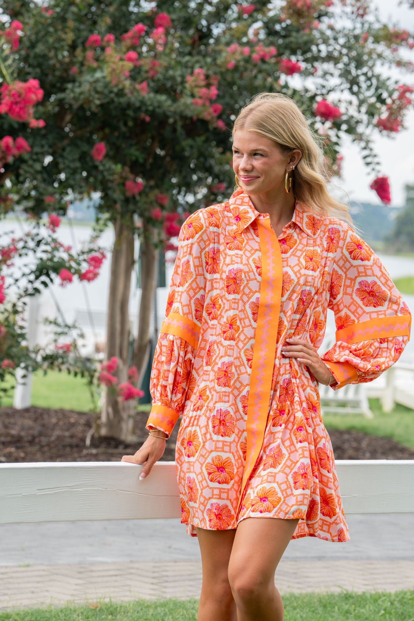 Wearing a cheerful Delany Dress - Floral Crochet with vibrant orange and pink motifs, an individual stands outdoors, smiling. They are near a wooden fence surrounded by blooming pink trees with a body of water visible in the background.